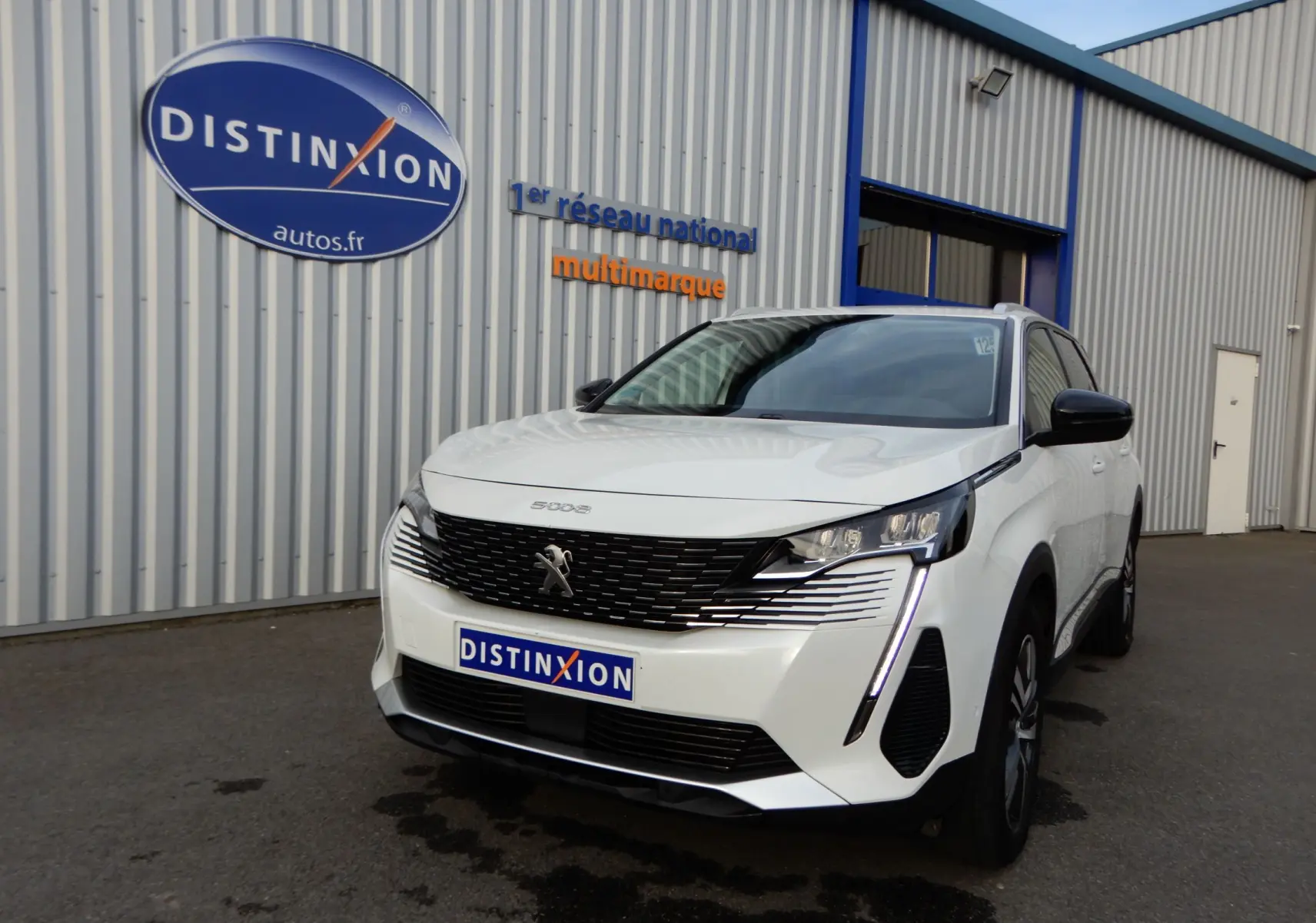 Peugeot 5008 blanc nacré en 3/4 avant droit, avec calandre noire et feux LED distinctifs, devant un bâtiment industriel.