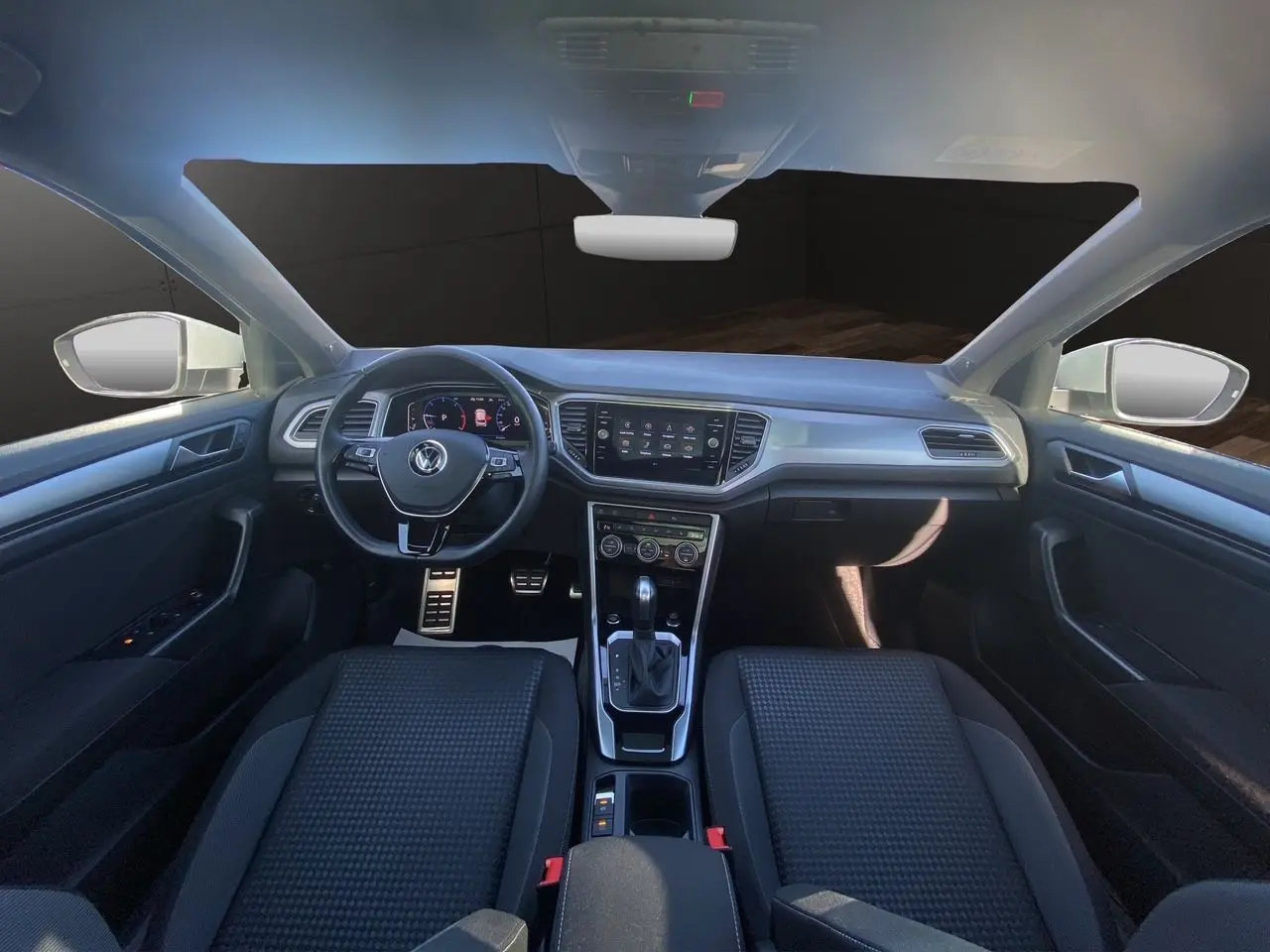 Vue intérieure avant du Volkswagen T-Roc 2021 avec tableau de bord noir, volant multifonction et toit ouvrant panoramique.