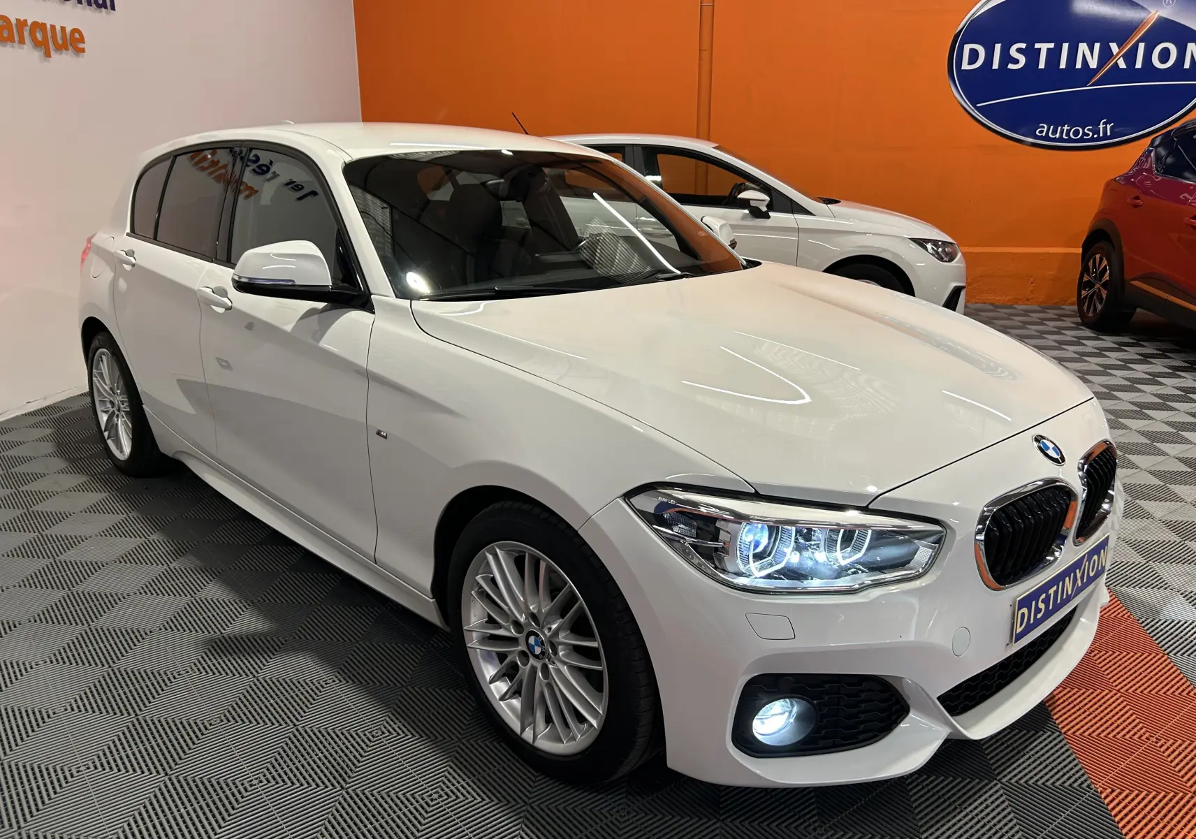 BMW Série 1 120iA blanc vue 3/4 avant droit avec phares allumés dans un showroom intérieur.