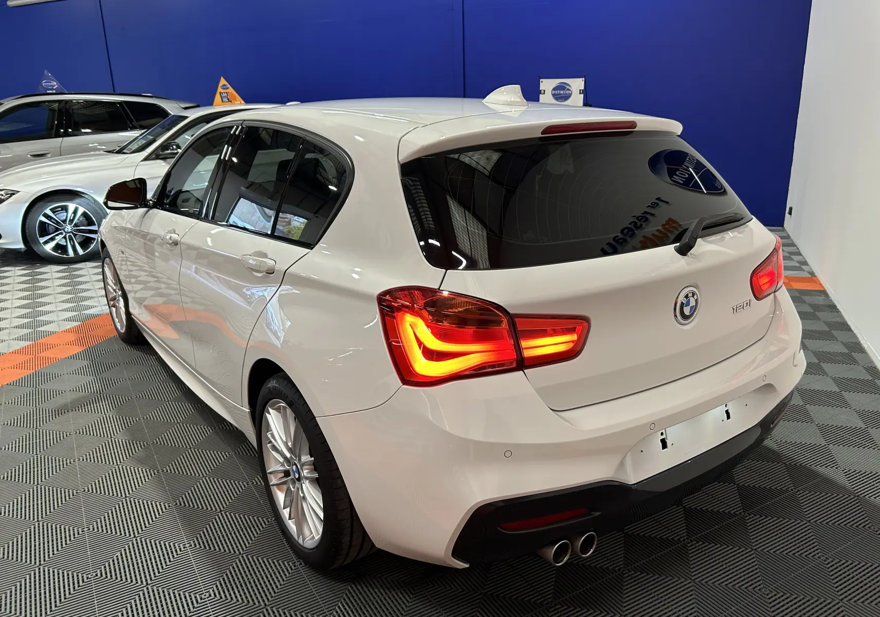 BMW Série 1 120iA M Sport blanche vue 3/4 arrière droit avec feux allumés dans un showroom.