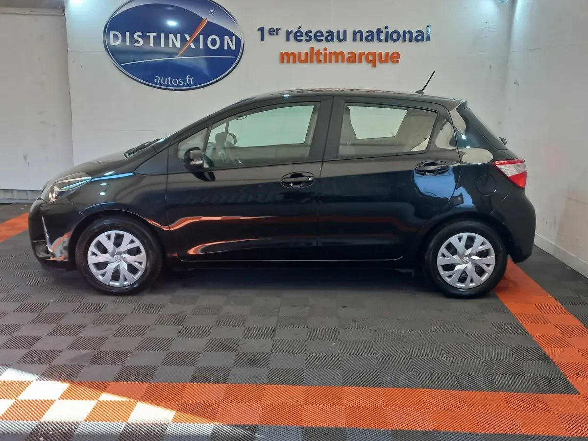 Profil côté gauche d'une Toyota Yaris noire hybride 2018 stationnée en intérieur avec jantes argentées et logo Distinxion en arrière-plan.