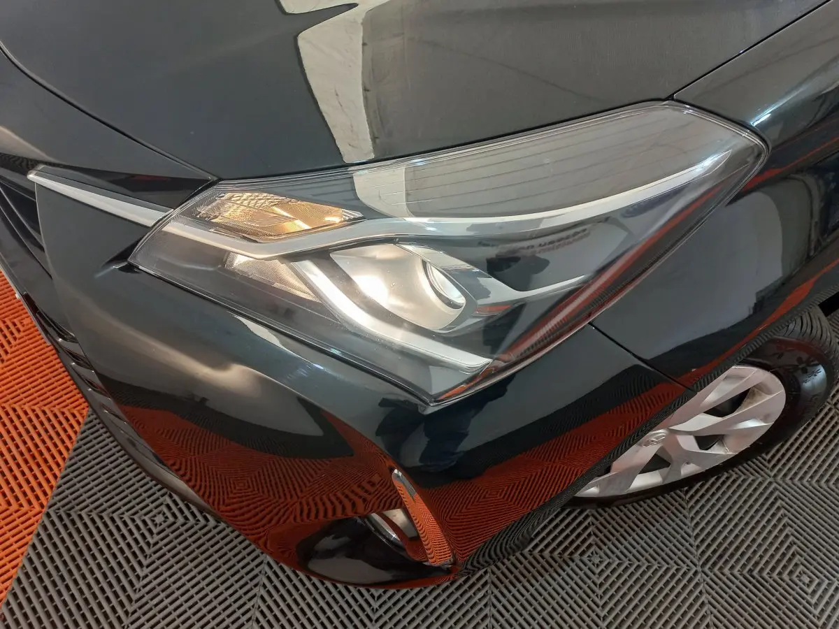 Gros plan sur le phare avant gauche d'une Toyota Yaris noire, avec partie de la roue et pare-chocs visibles.