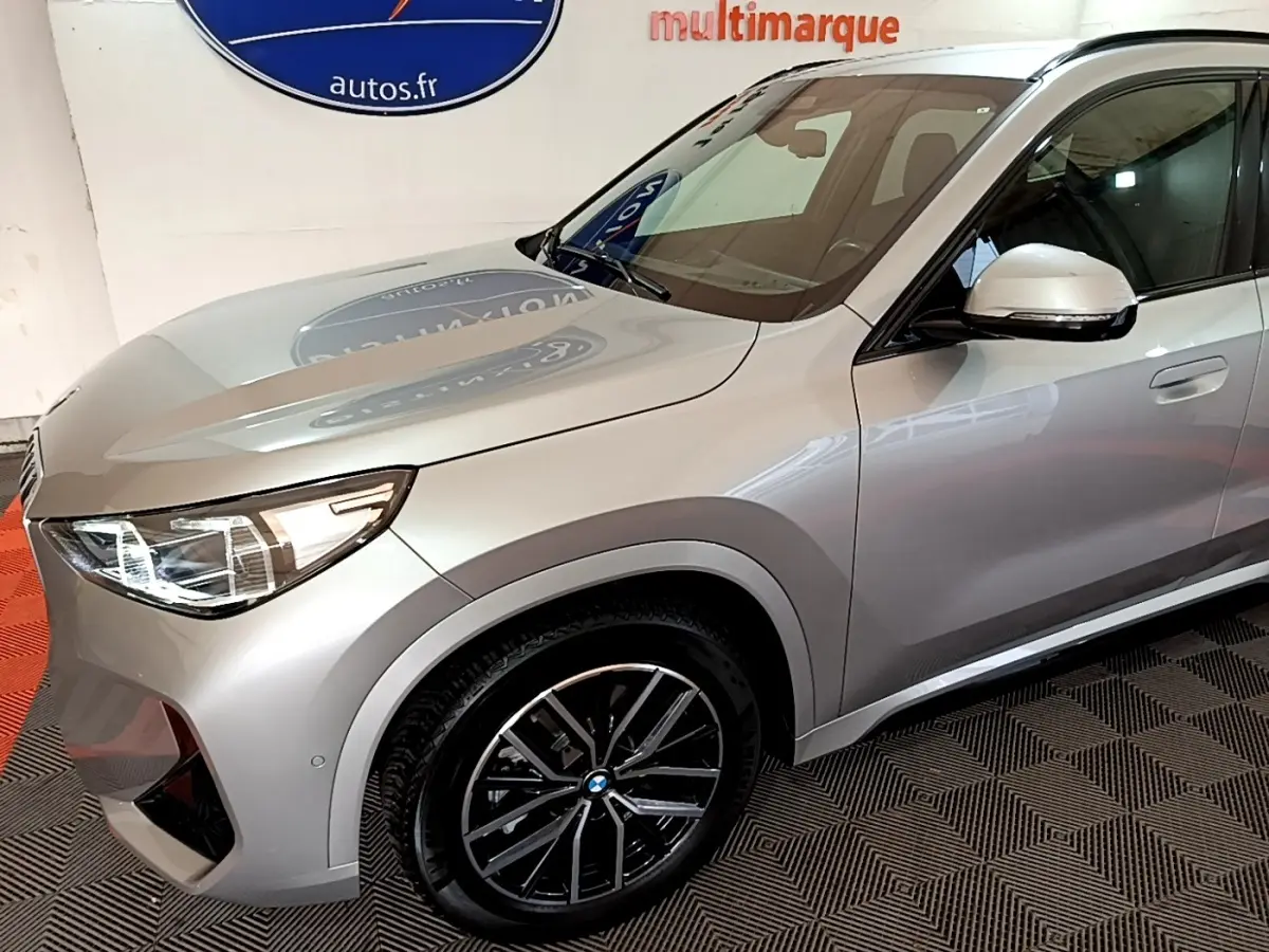 Vue 3/4 avant droit d’un BMW X1 xDrive23i M Sport gris clair avec jantes noires et phares LED allumés.