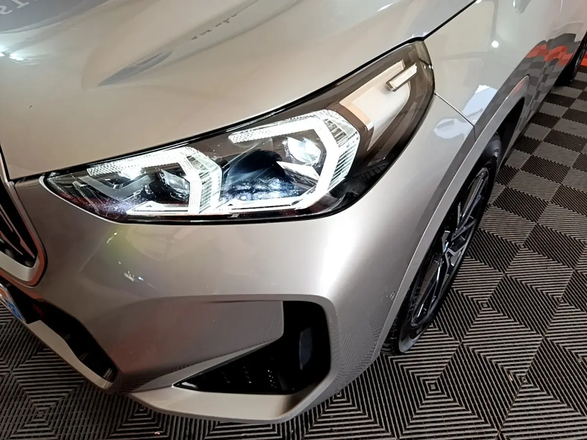 Gros plan sur l'avant droit gris clair du BMW X1 xDrive23i M Sport 2023, mettant en valeur le phare LED et la jante noire.