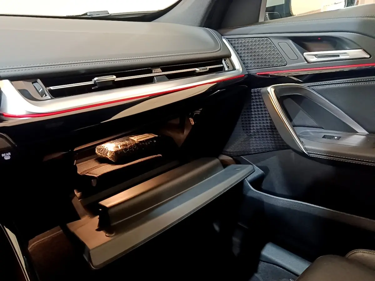 Vue intérieure côté passager du BMW X1 xDrive23i M Sport 2023, tableau de bord noir avec éclairage d'ambiance rouge et boîte à gants ouverte.