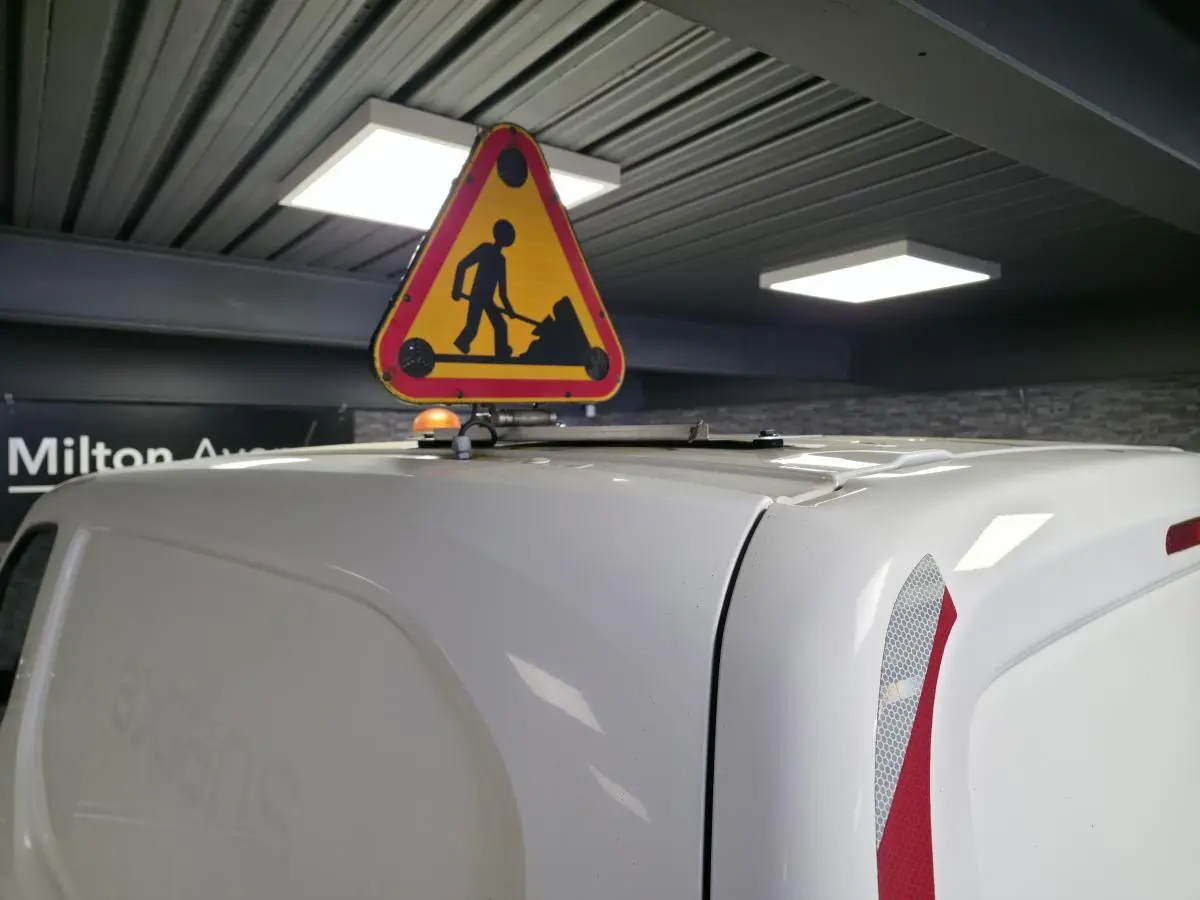 Vue arrière droite d’un Peugeot Partner blanc avec panneau de signalisation travaux fixé sur le toit en intérieur.