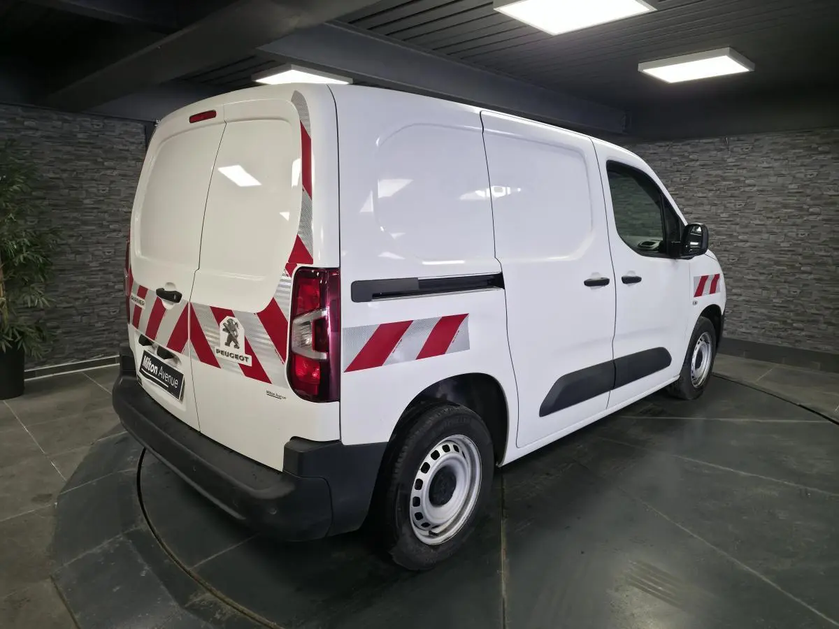 Vue 3/4 arrière droite d’un Peugeot Partner blanc avec bandes réfléchissantes rouges et protections noires.