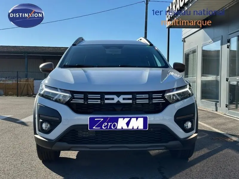Vue frontale d'un Dacia Jogger blanc avec calandre noire et phares LED allumés sur parking extérieur.