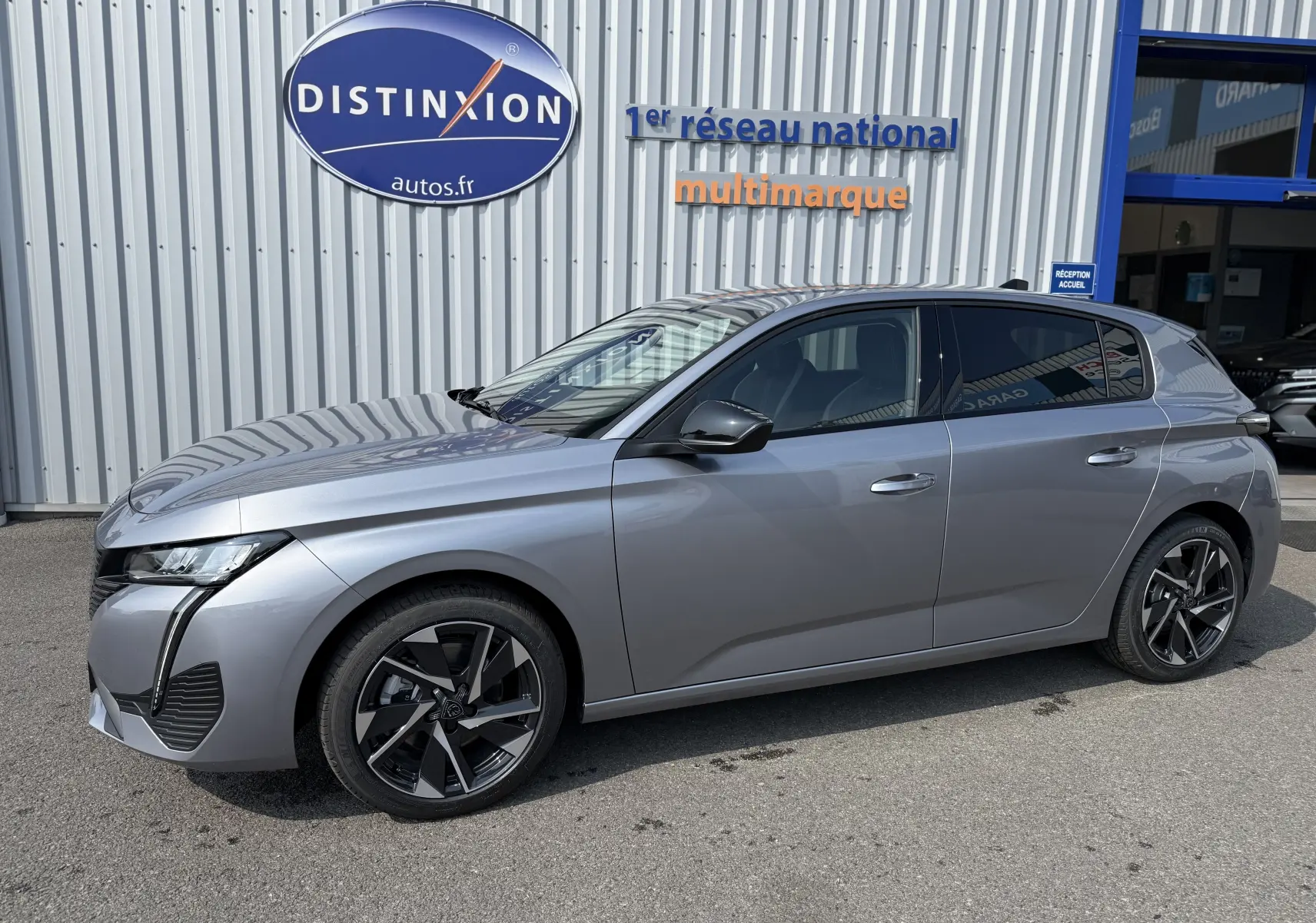 Peugeot 308 gris Artense vue de profil côté gauche, avec jantes noires et vitres teintées, devant un garage multimarque.