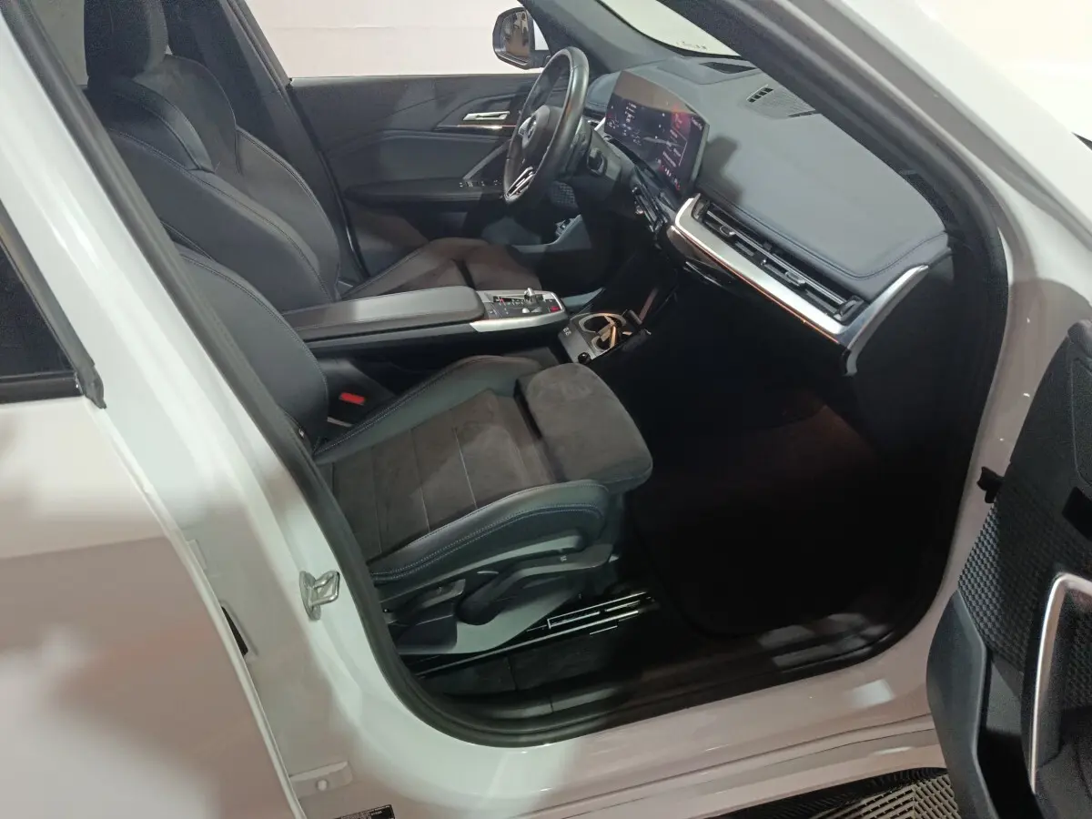 Vue intérieure côté conducteur d'une BMW X1 blanche 2024, avec sièges noirs et tableau de bord digital lumineux.