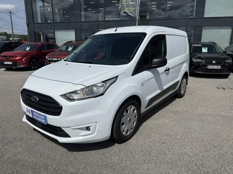 Ford Transit Connect blanc glacier vu en 3/4 avant droit sur parking devant un bâtiment vitré.