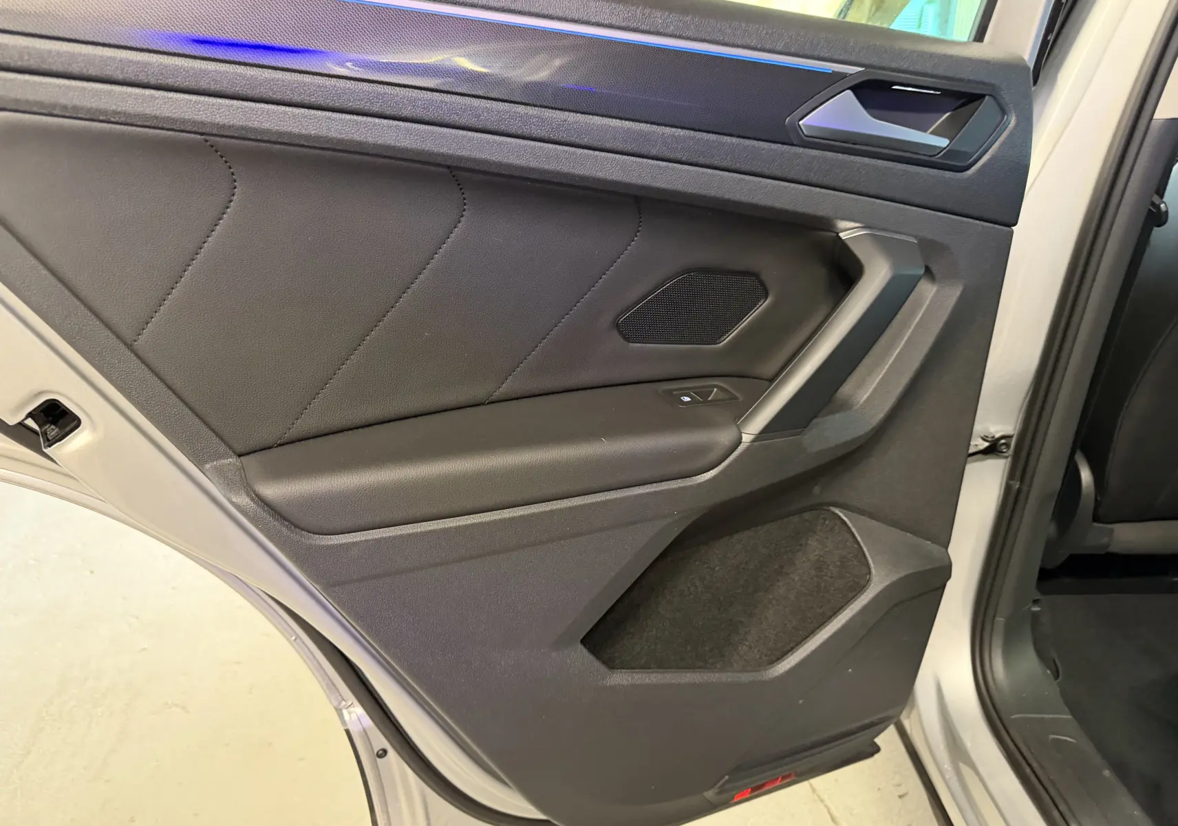 Vue intérieure de la porte arrière gauche gris clair du Volkswagen Tiguan 1.4 E-HYBRID avec garniture noire et éclairage d'ambiance bleu.