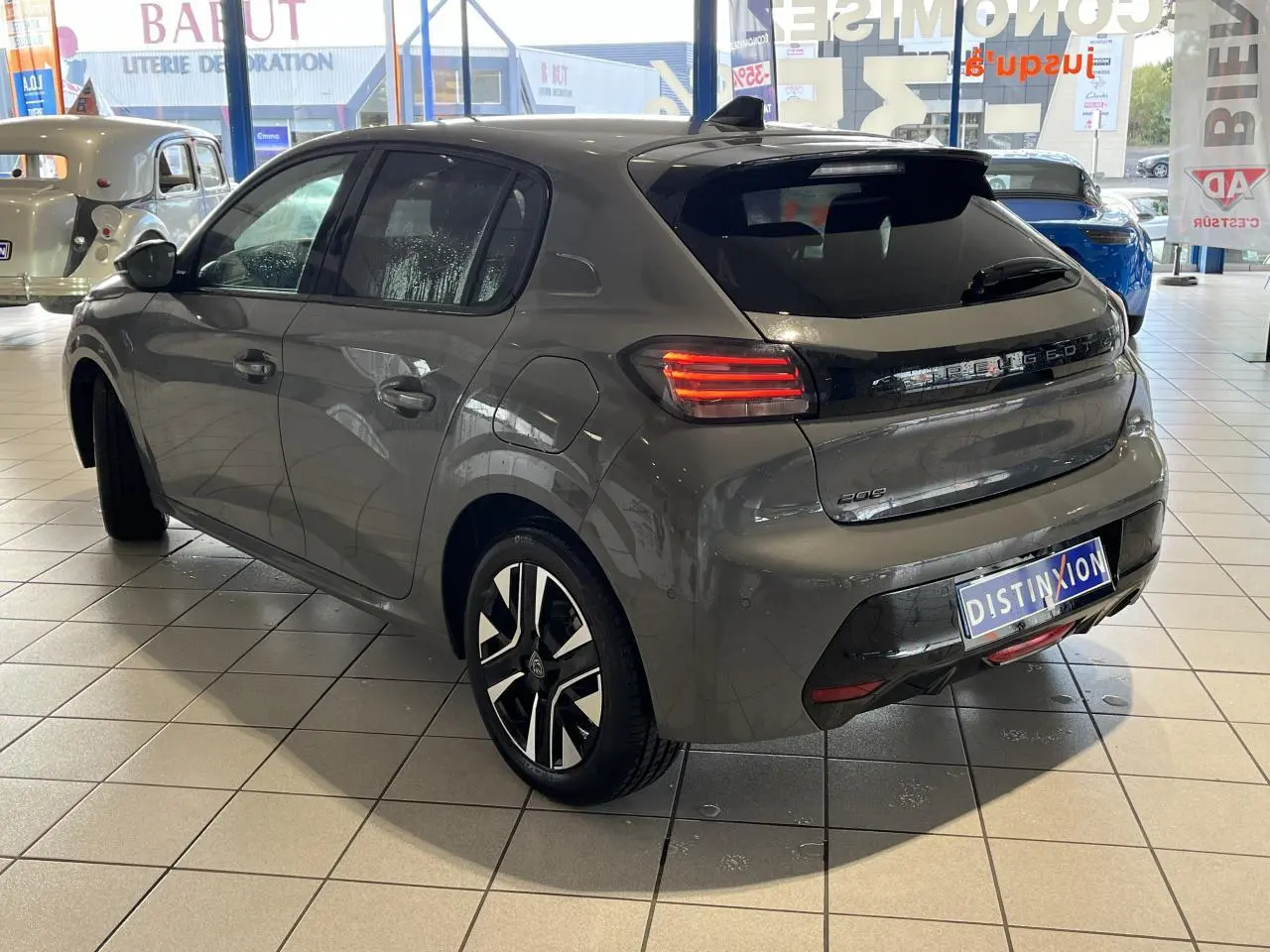 Vue 3/4 arrière droite de la Peugeot 208 2024 gris clair Selenium avec feux LED et jantes alliage bicolores en showroom.