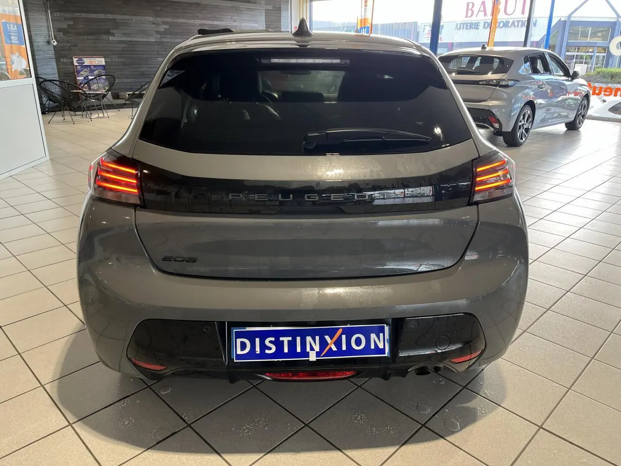Vue arrière d'une Peugeot 208 gris clair Selenium avec bandeau noir brillant et feux arrière à LED en intérieur de showroom.