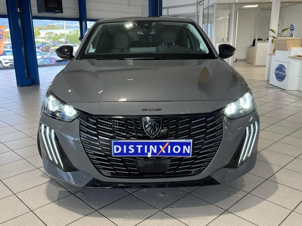 Vue avant d'une Peugeot 208 2024 gris clair Selenium avec calandre noire et feux LED allumés en showroom.