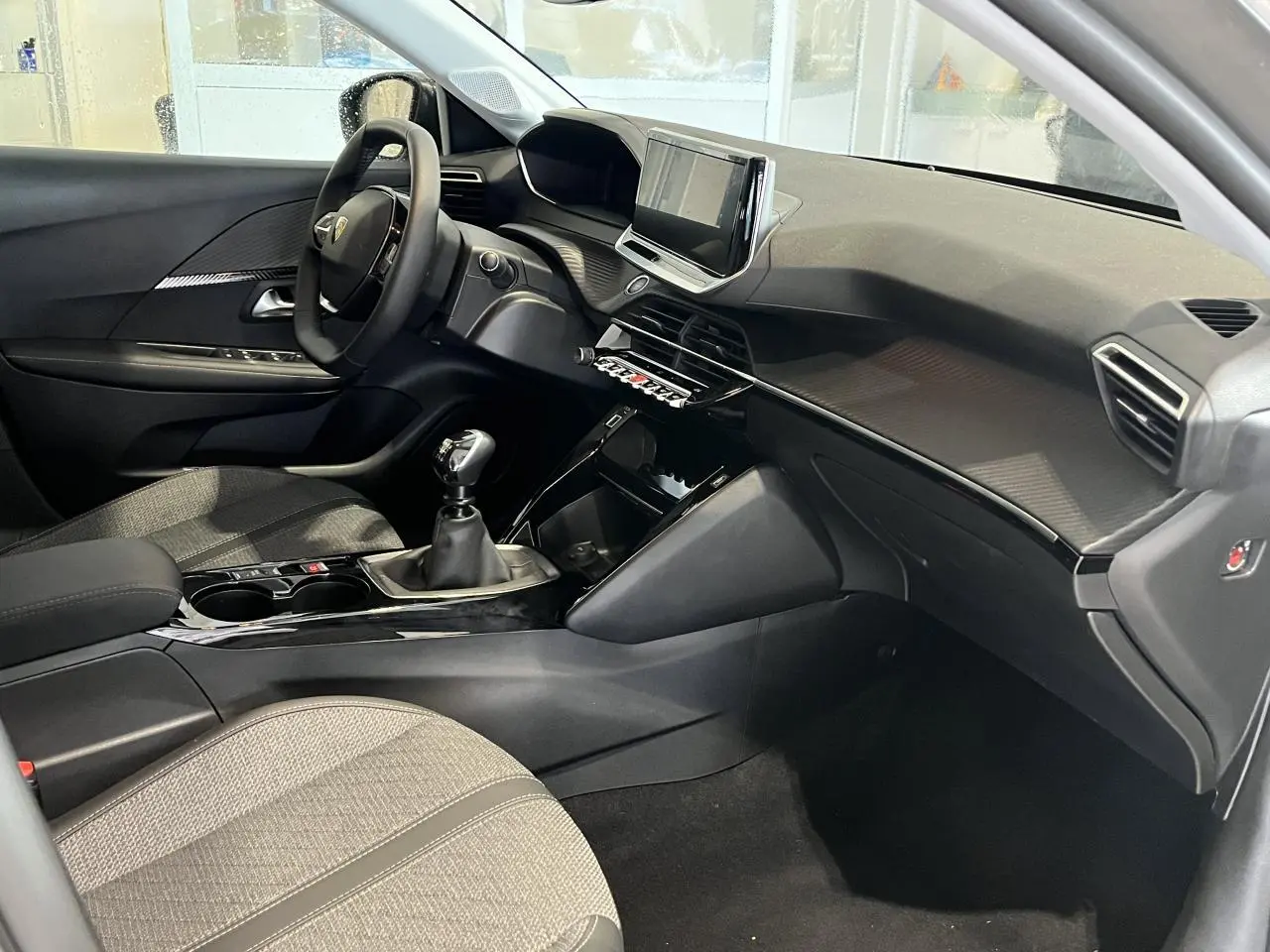 Vue intérieure côté conducteur de la Peugeot 208 2024, tableau de bord noir avec écran tactile et sellerie tissu gris clair.