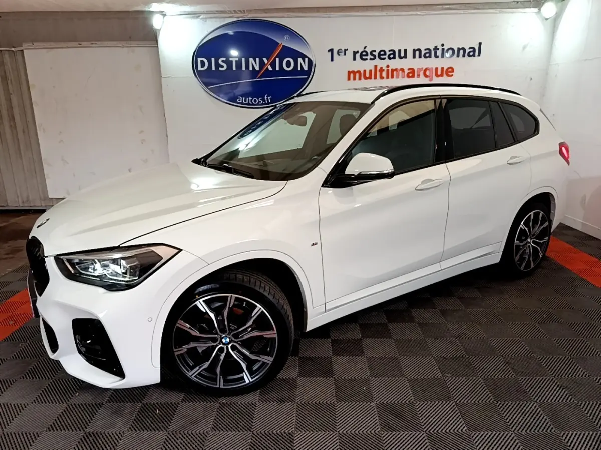 BMW X1 sDrive18i M Sport blanc vue 3/4 avant droit, jantes alliage noires et phares LED allumés, en intérieur showroom.