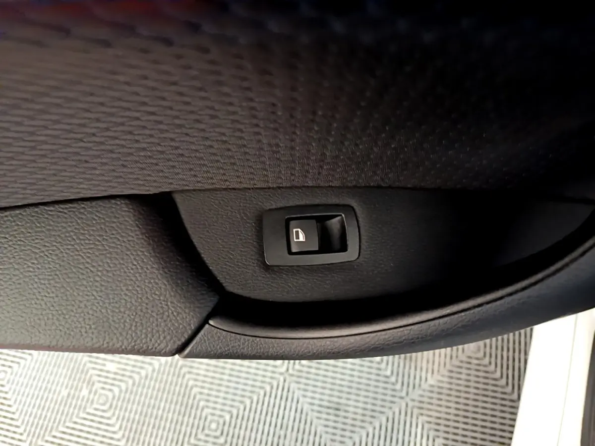 Gros plan sur le bouton de fermeture électrique du coffre, intégré dans la garniture noire de la porte d’un BMW X1 blanc.