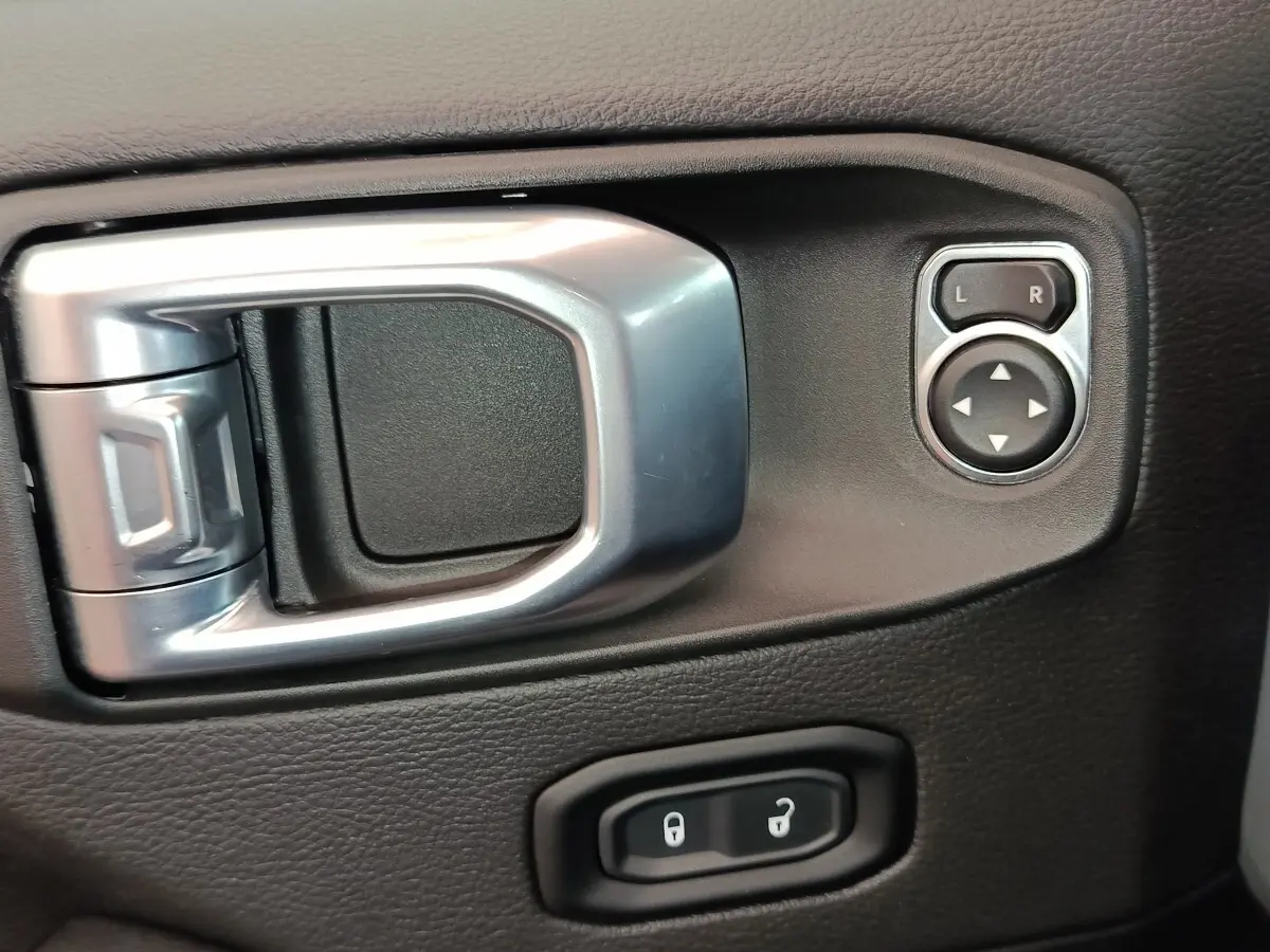 Gros plan sur la poignée intérieure argentée et les commandes de réglage des rétroviseurs du côté conducteur d’un Jeep Wrangler gris foncé.