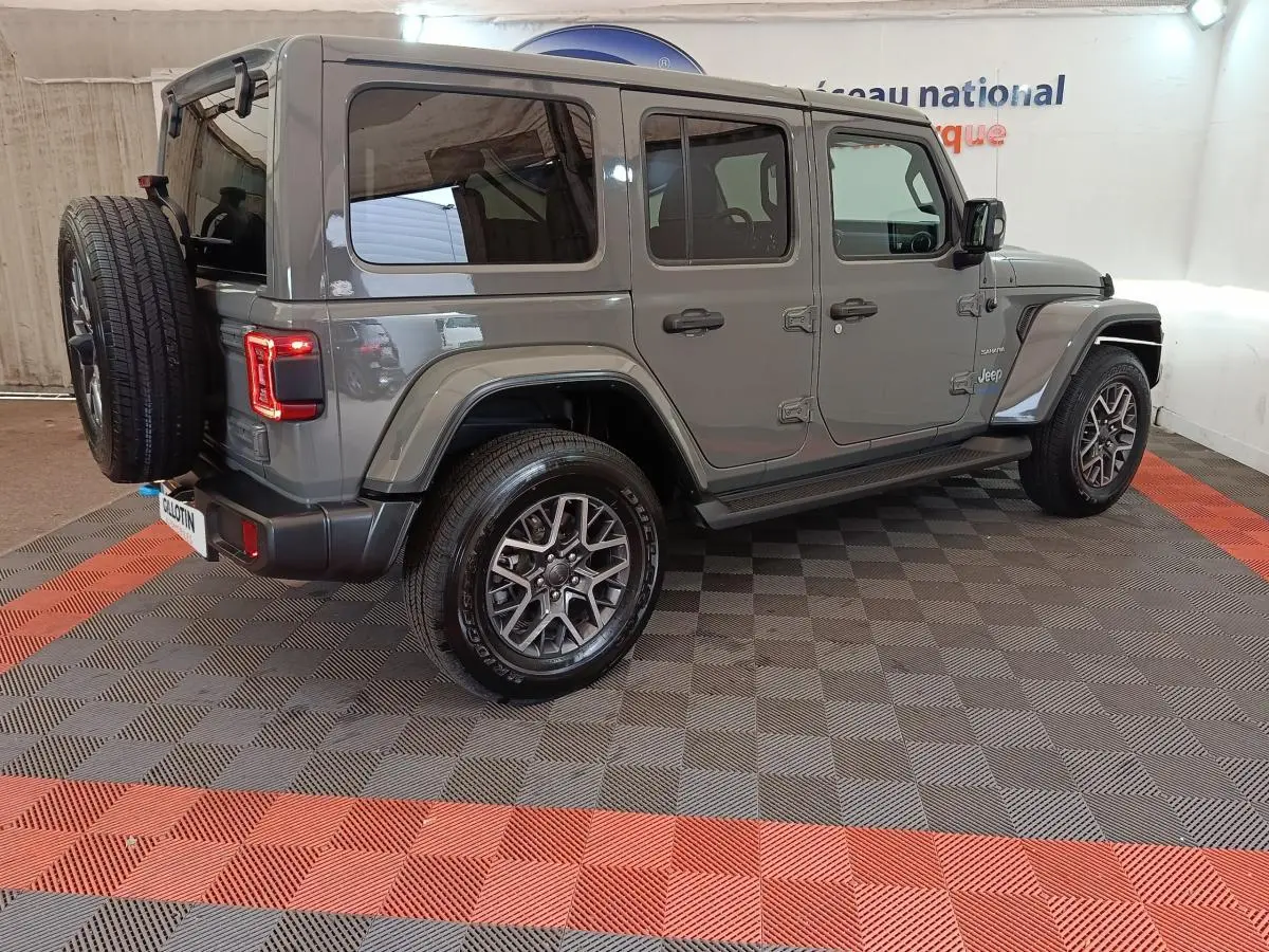 Jeep Wrangler gris foncé vue 3/4 arrière droit, avec roue de secours montée à l'arrière et feux LED allumés.
