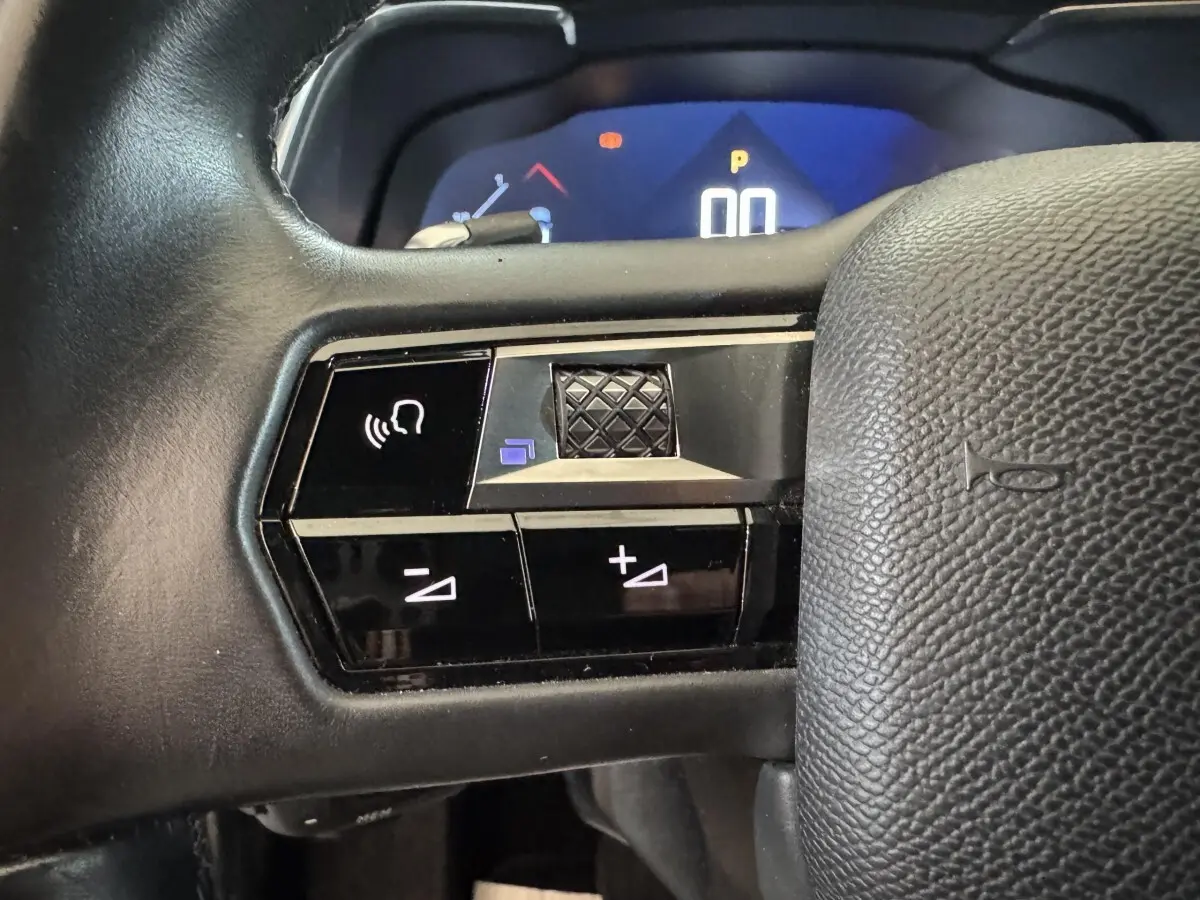 Gros plan sur les commandes vocales et volume au volant cuir noir du DS7 Crossback gris foncé, tableau de bord numérique en arrière-plan.