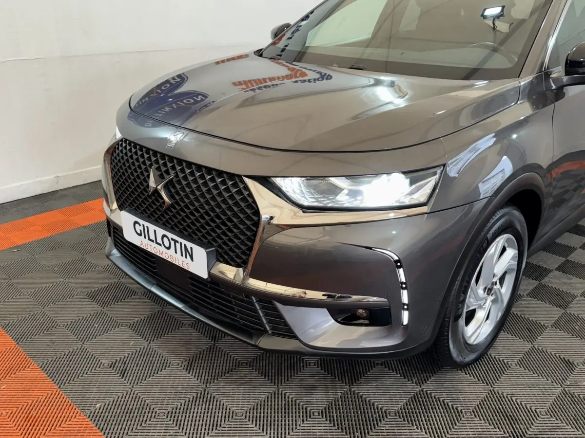 Vue 3/4 avant droit du DS7 Crossback gris foncé avec calandre distinctive et feux LED allumés en intérieur showroom.