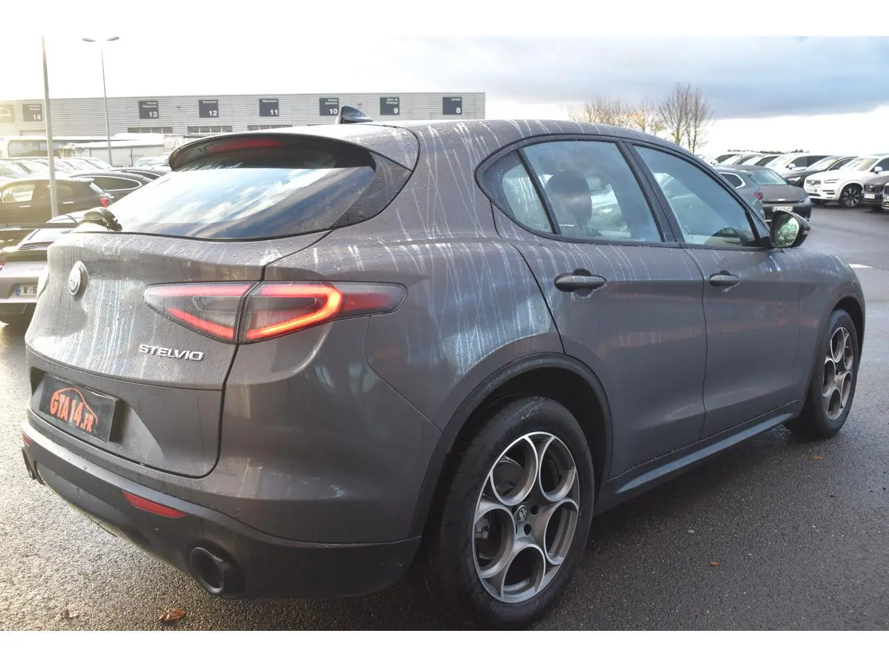 Vue 3/4 arrière droite d'un Alfa Romeo Stelvio 2024 gris Vesuvio avec jantes 19'' et feux arrière allumés.