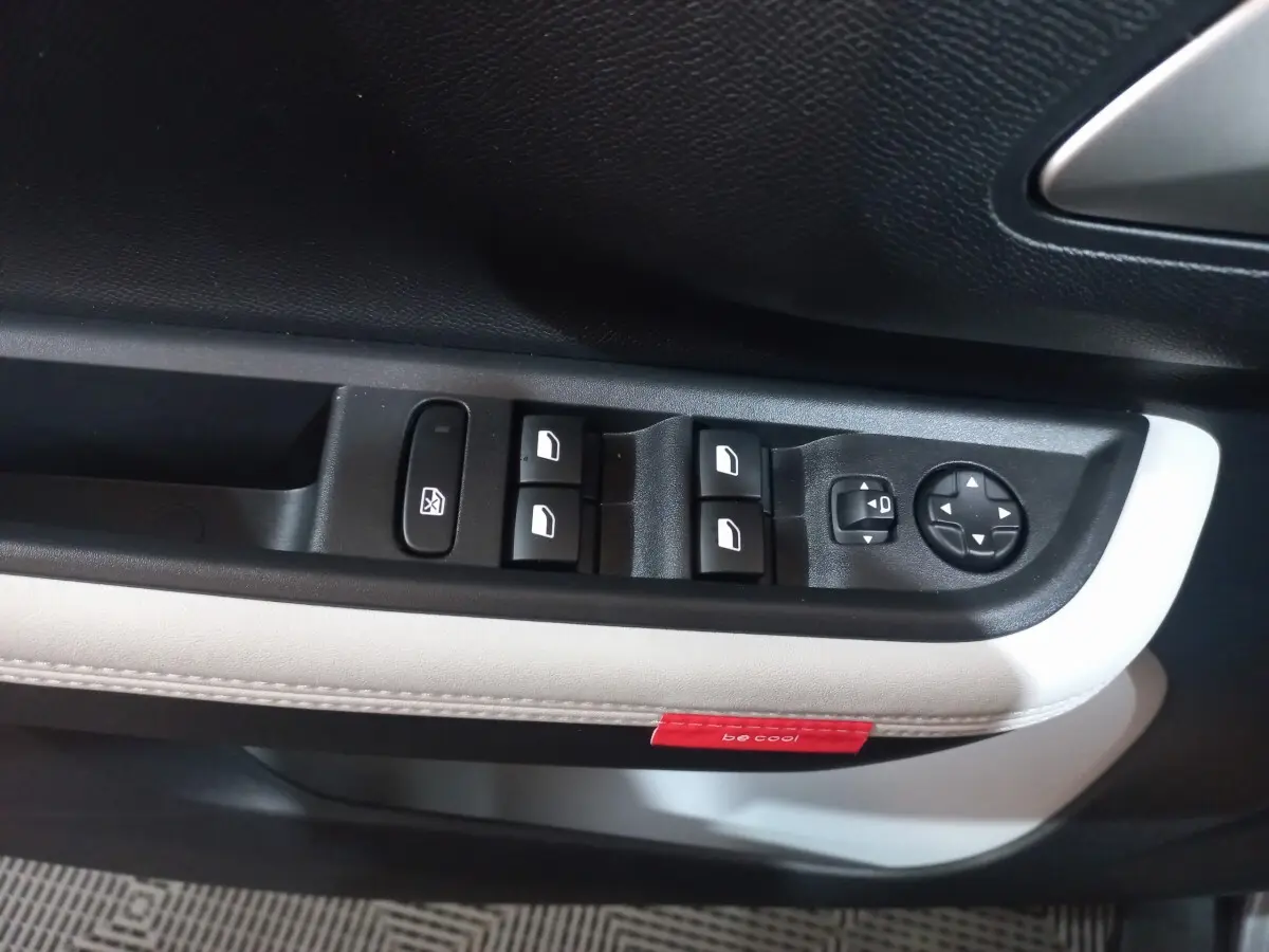 Gros plan sur la commande des vitres et rétroviseurs côté conducteur de la Citroën C3 hybride gris foncé avec surpiqûres blanches et étiquette rouge "be cool".