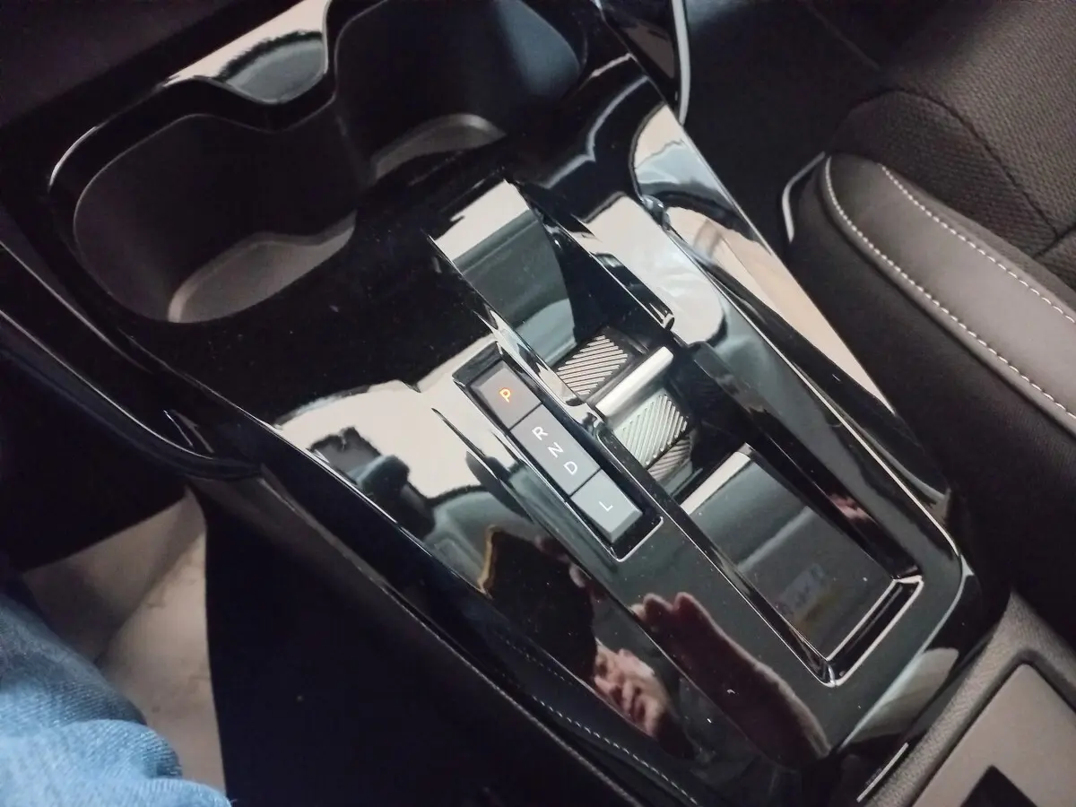 Gros plan sur la console centrale noire brillante avec levier de vitesses automatique et porte-gobelets, siège tissu gris visible.