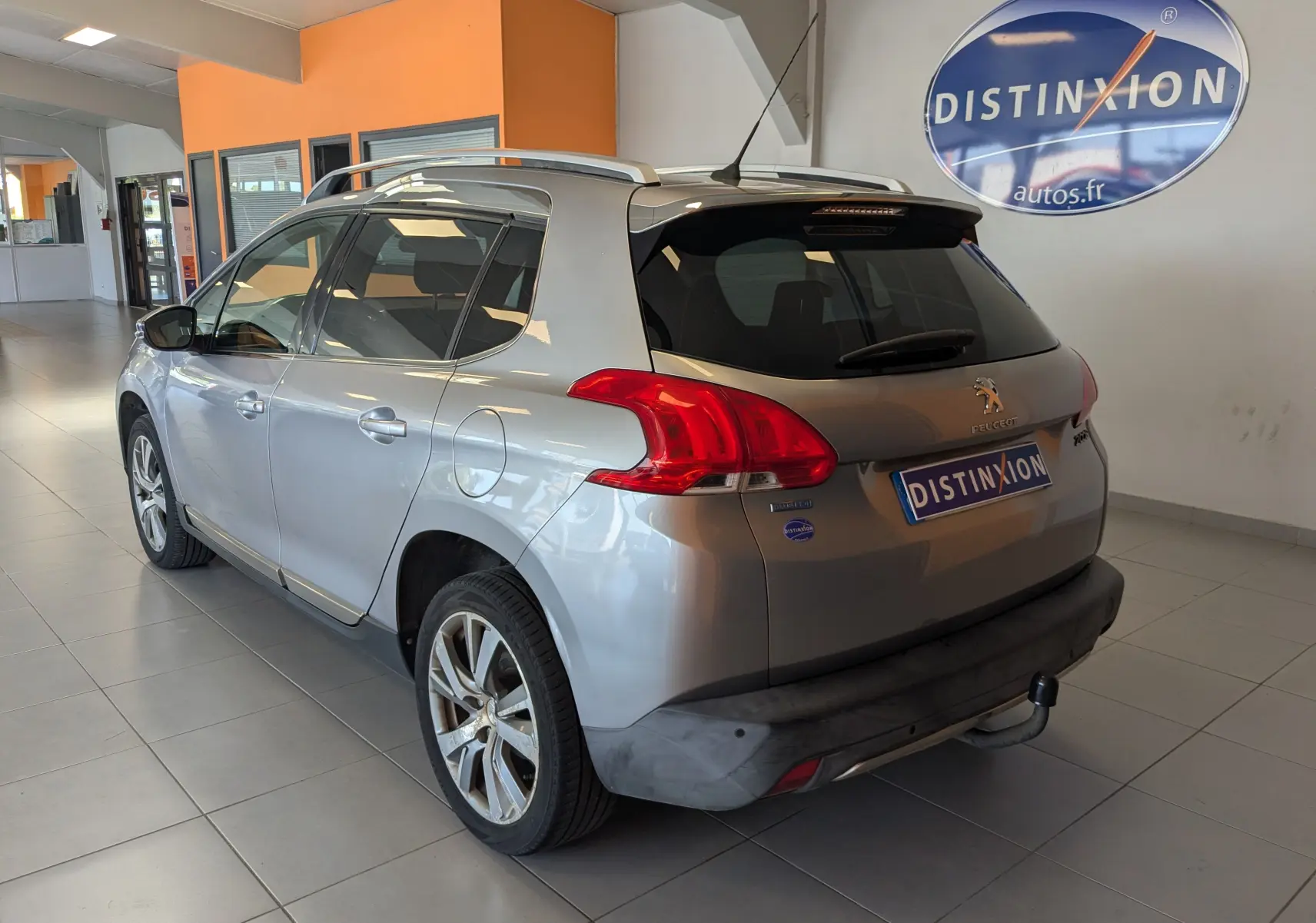 Vue 3/4 arrière droite d’un Peugeot 2008 gris Artense en showroom, avec barres de toit et attelage visible.
