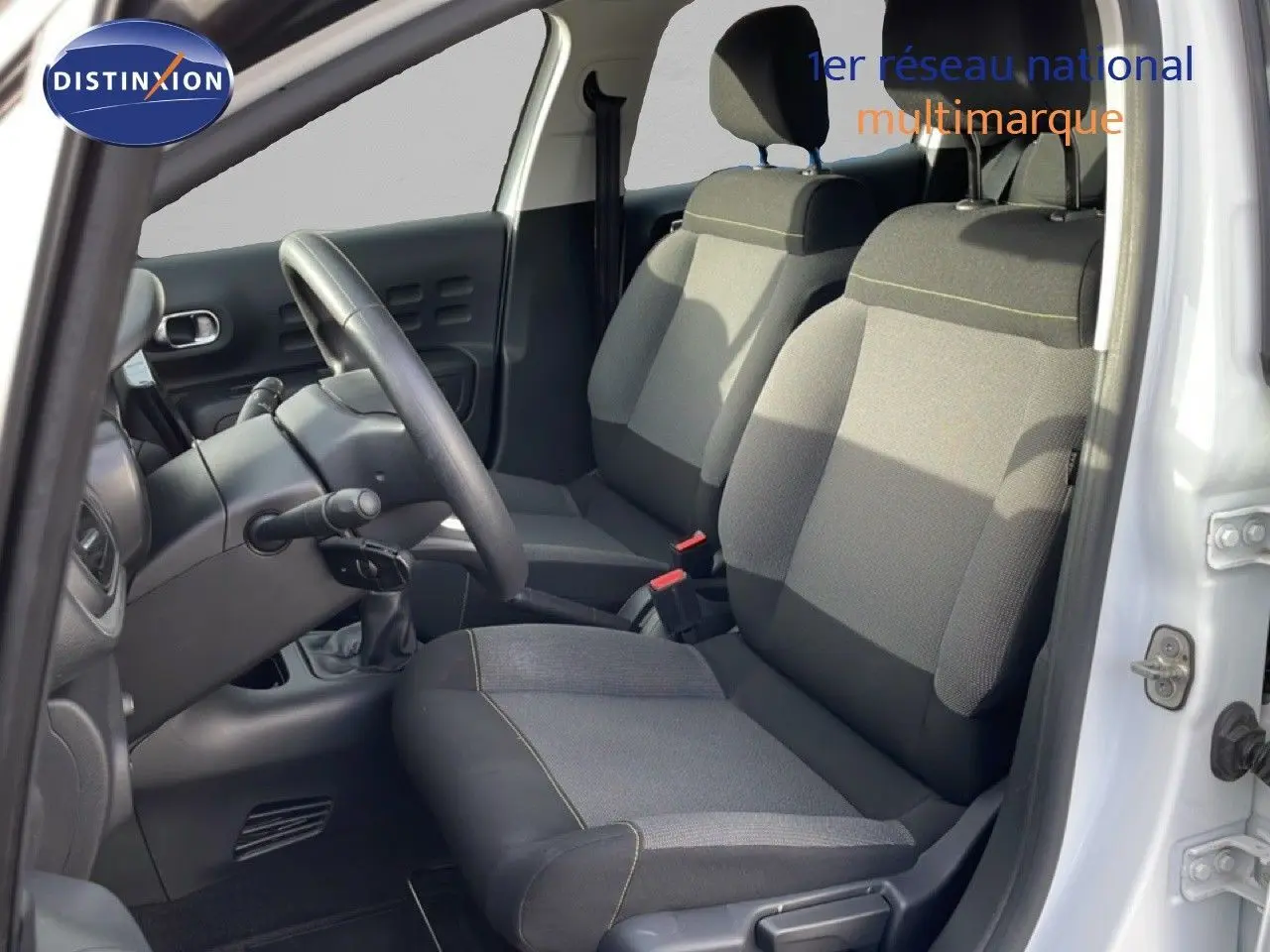 Vue intérieure côté conducteur de la Citroën C3 blanche avec toit rouge, montrant les sièges avant en tissu gris et volant noir.