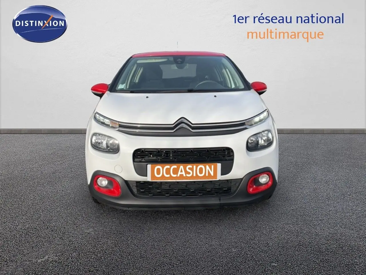 Vue frontale d'une Citroën C3 blanche avec toit et détails rouges, sur fond neutre avec panneau publicitaire.