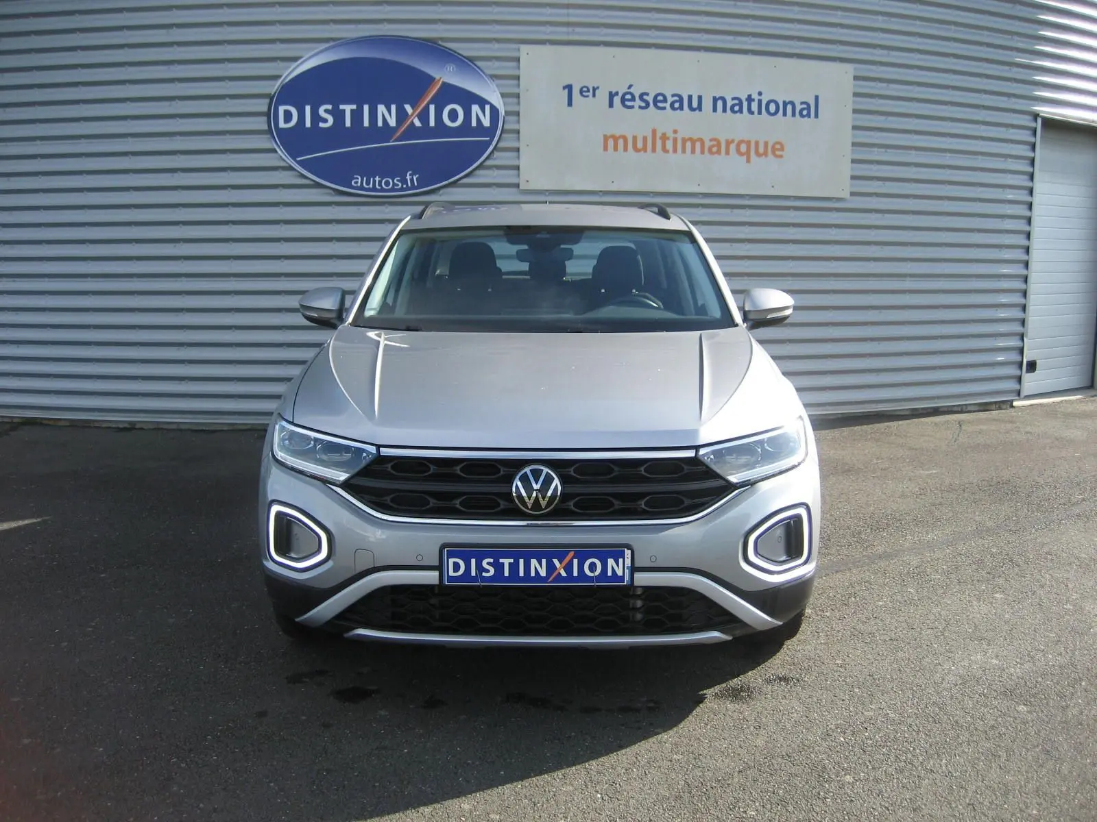 Vue frontale d'un Volkswagen T-Roc gris clair 2023 avec feux LED et calandre noire, devant un mur métallique.