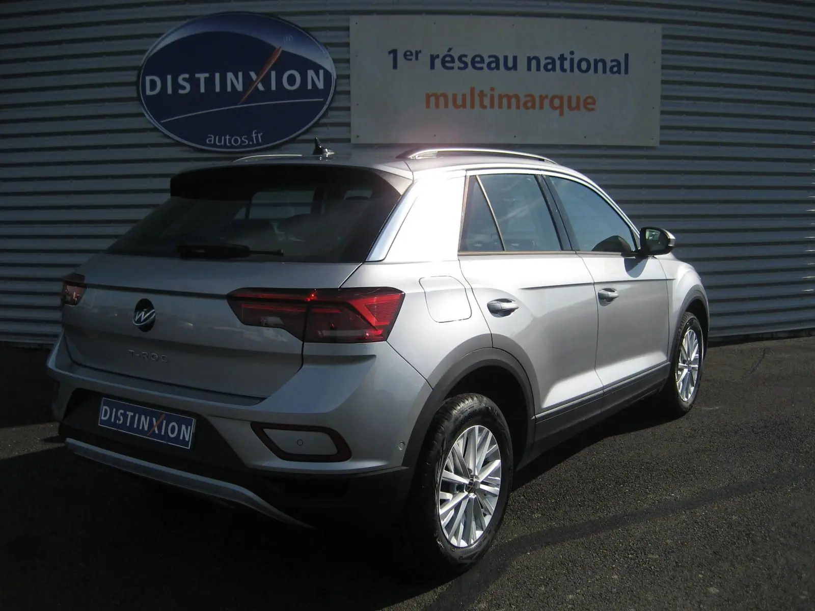 Vue 3/4 arrière droite d’un Volkswagen T-Roc gris clair 2023 garé devant un mur métallique avec logo Distinxion.