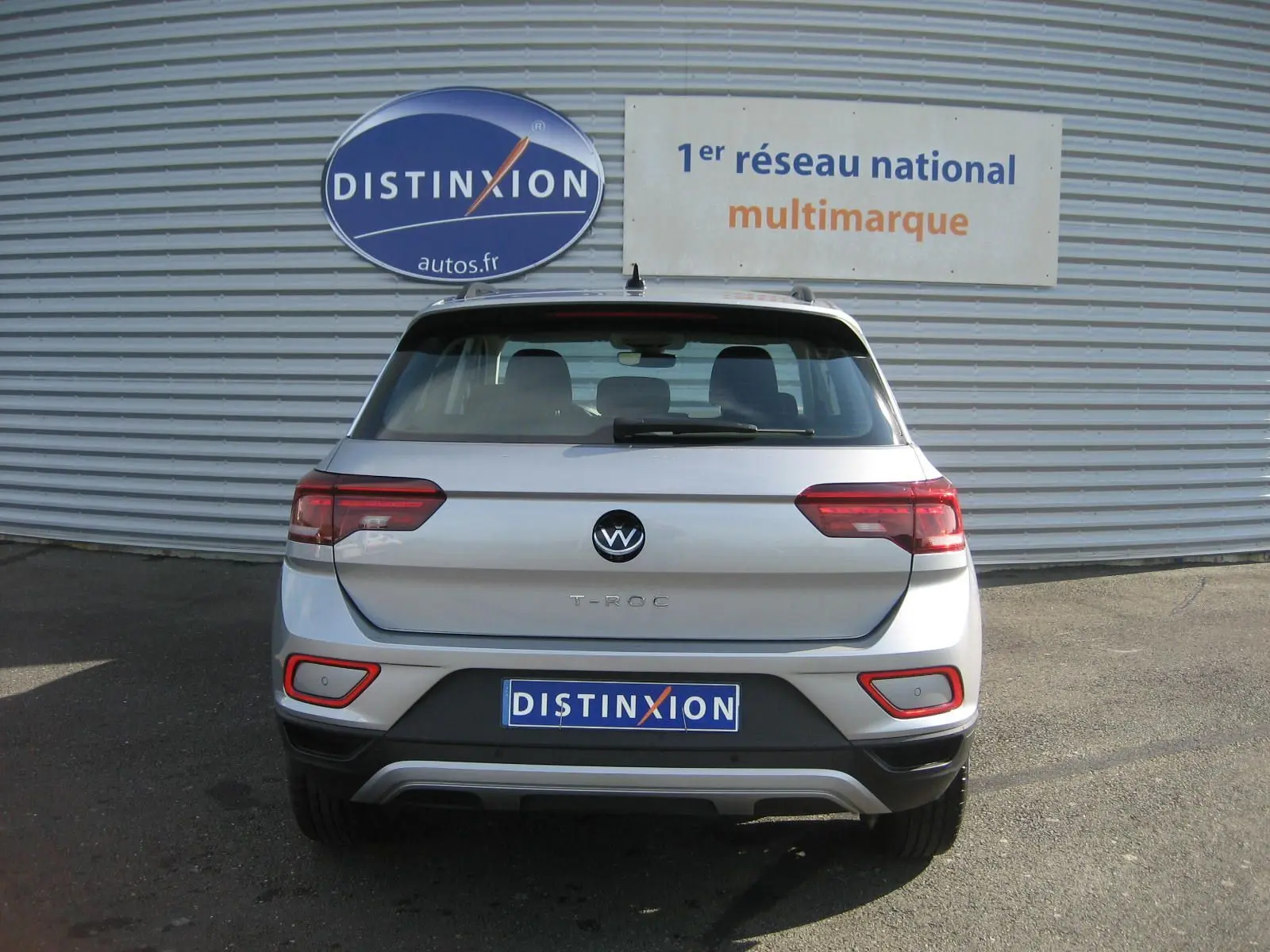 Vue arrière d’un Volkswagen T-Roc gris clair 2023 avec feux LED et logo distinctif sur fond de façade métallique.