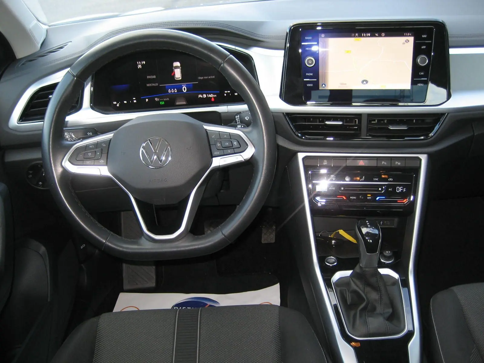 Vue intérieure frontale du volant et tableau de bord du Volkswagen T-Roc gris clair avec écran tactile et levier de boîte automatique.