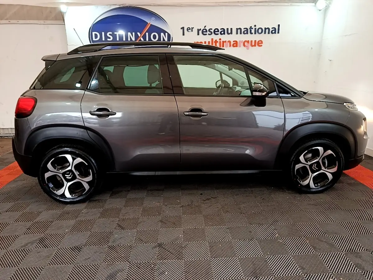 Vue de profil côté gauche d’un Citroën C3 Aircross gris foncé avec barres de toit noires en intérieur showroom.