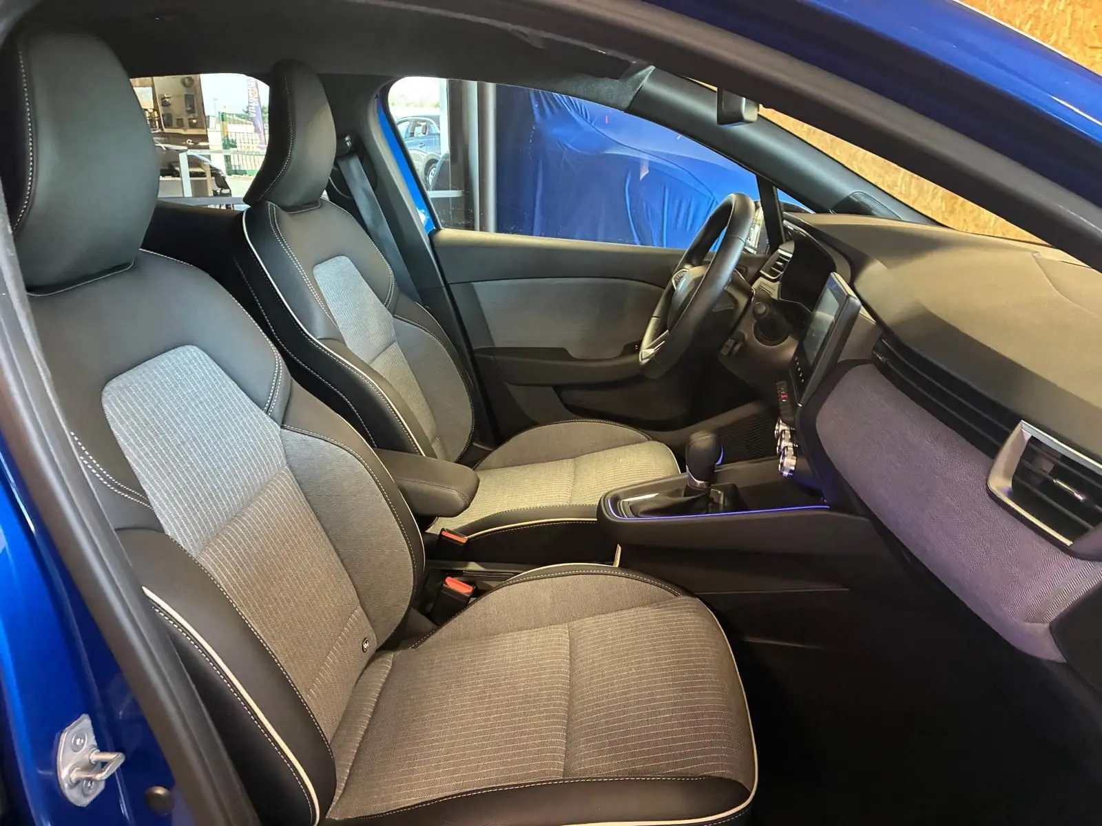 Vue intérieure côté conducteur de la Renault Clio V E-TECH 145 TECHNO 2025 bleu iron, sièges tissu gris et noir, tableau de bord moderne.