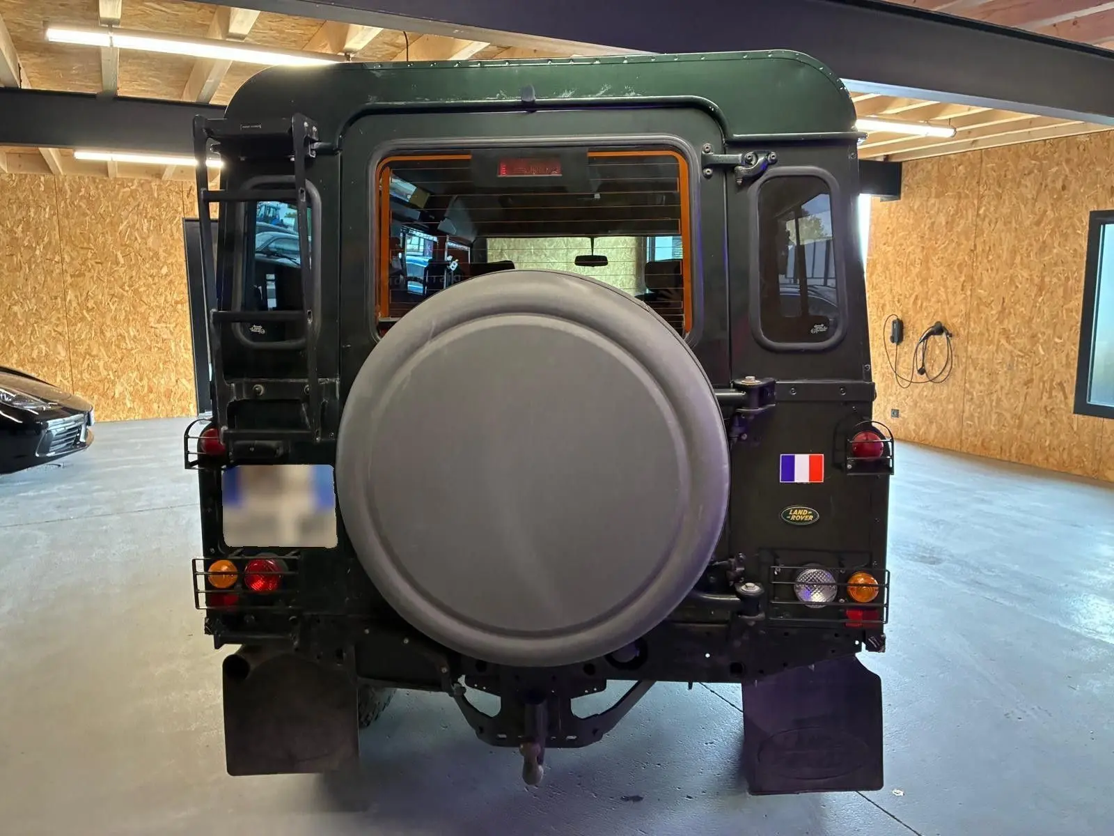 Vue arrière d’un Land Rover Defender 110 vert foncé de 2005, avec roue de secours et drapeau français sur la porte arrière.