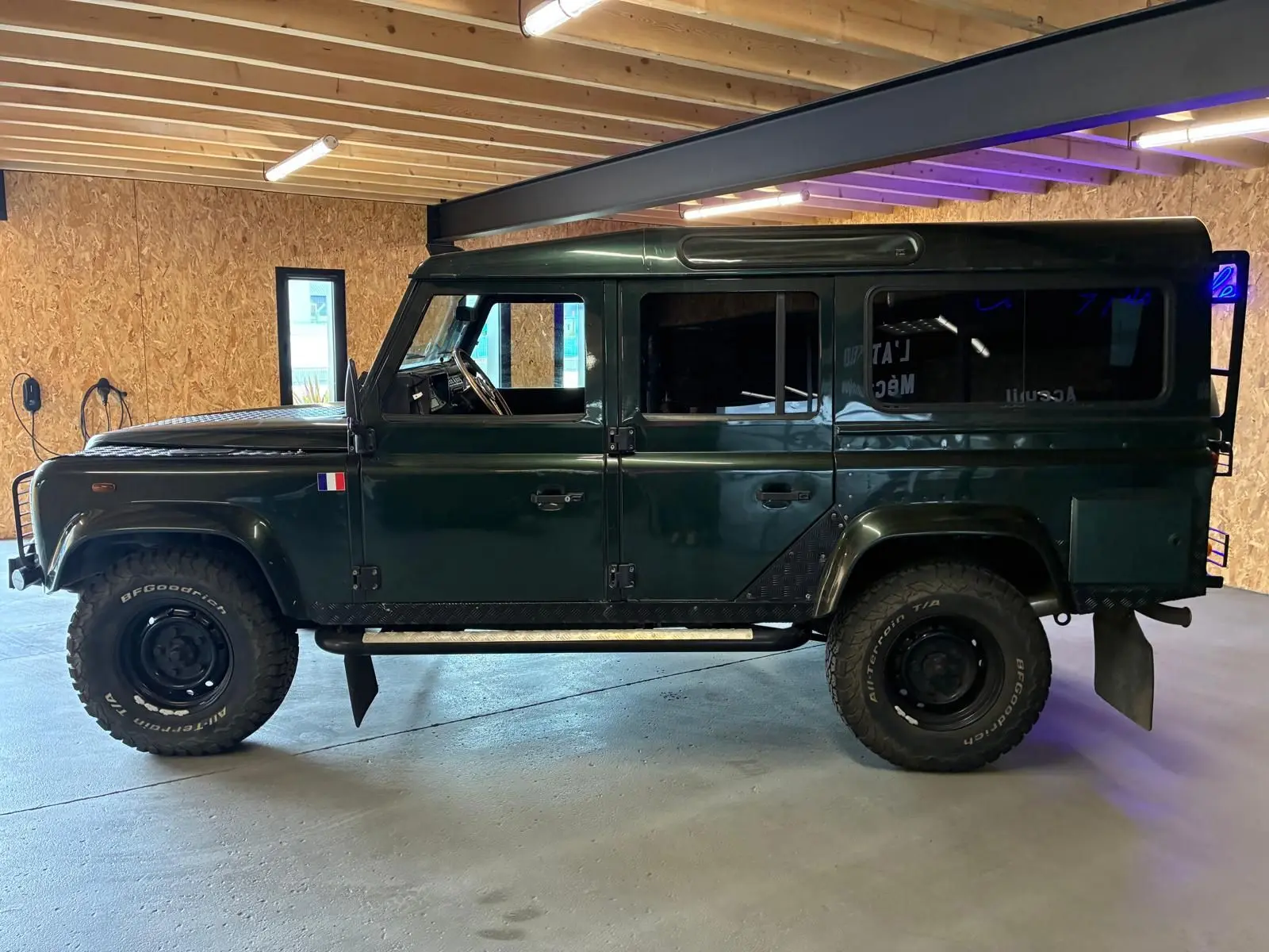 Vue de profil côté gauche d'un Land Rover Defender 110 vert foncé de 2005 avec pneus tout-terrain et drapeau français sur l'aile avant.