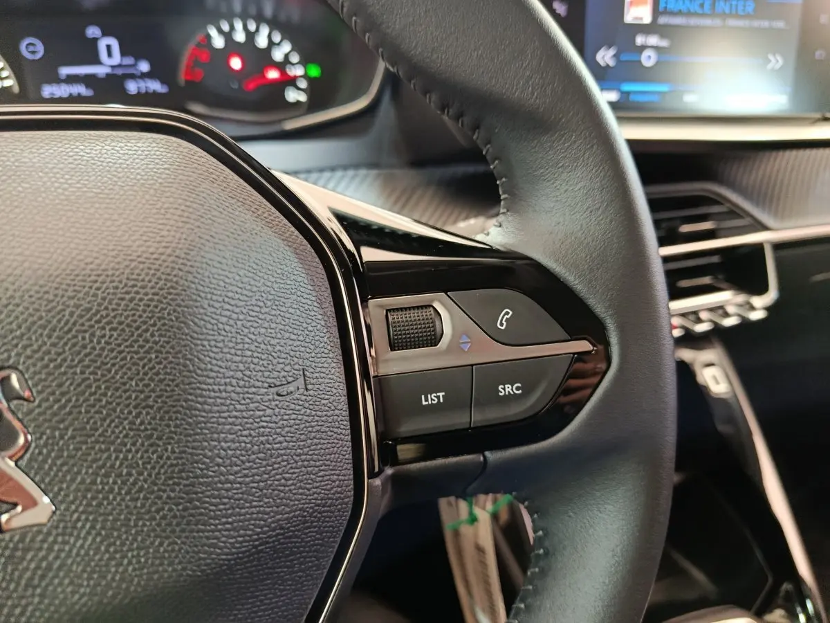 Gros plan sur le volant cuir multifonctions de la Peugeot 208 blanche, avec commandes audio et tableau de bord en arrière-plan.