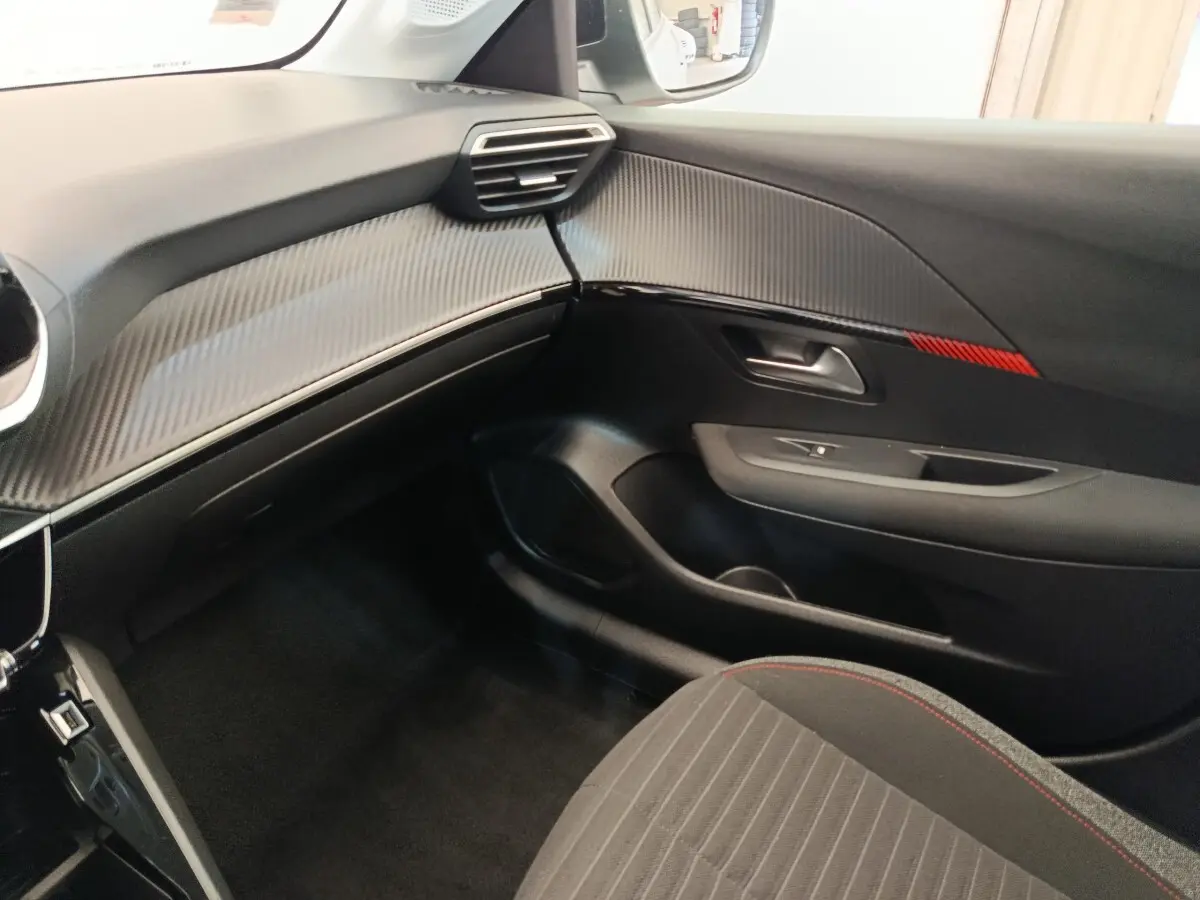 Vue intérieure côté passager de la Peugeot 208 blanche, tableau de bord avec insert carbone et bande rouge décorative.