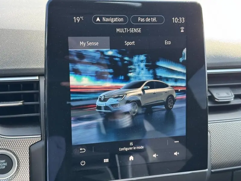 Écran tactile intérieur affichant une Renault Arkana gris clair en vue 3/4 avant sur fond urbain flou.