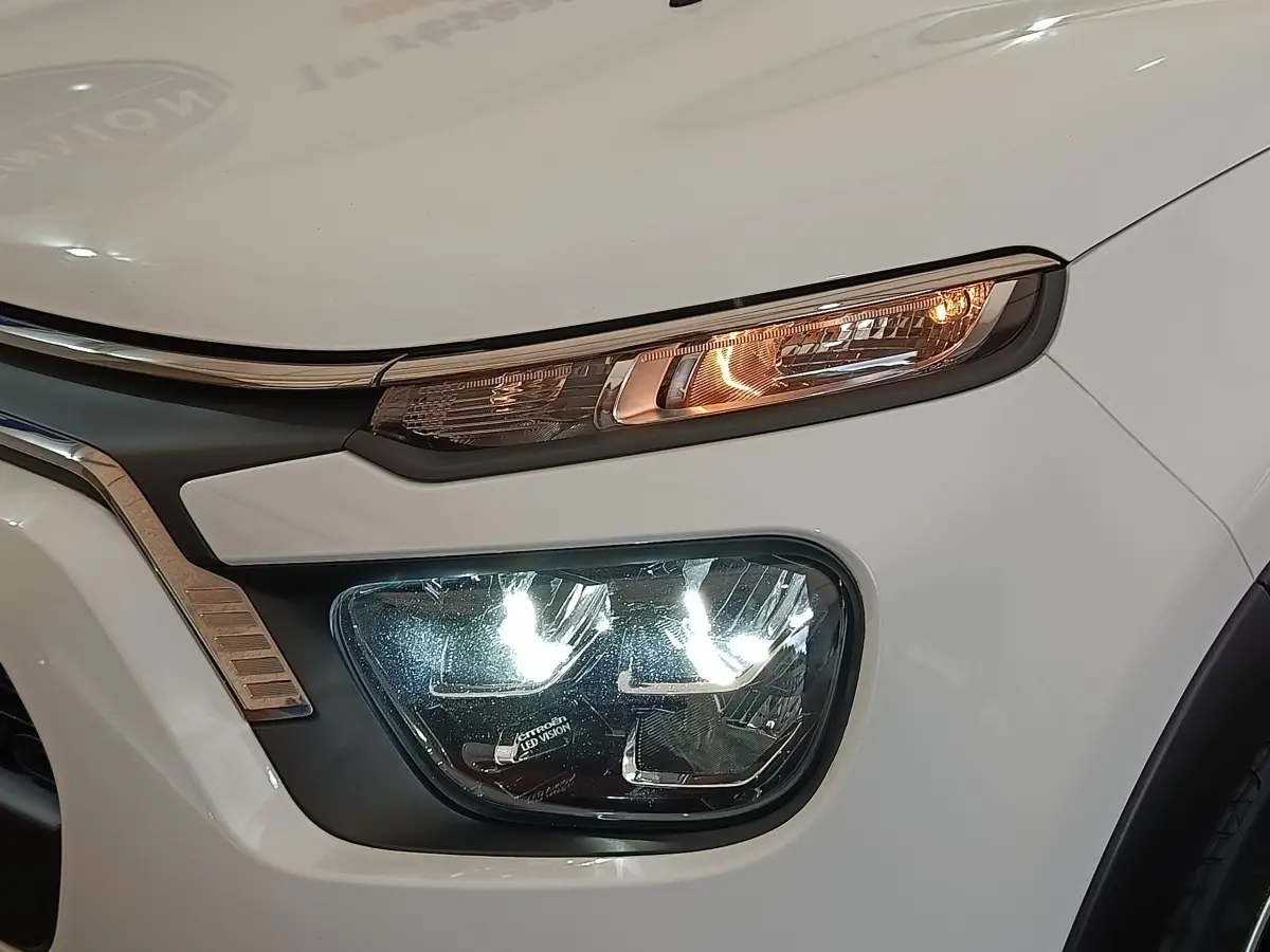 Gros plan sur l'avant droit blanc d'une Citroën C3 2022, mettant en valeur ses phares LED allumés et la calandre chromée.