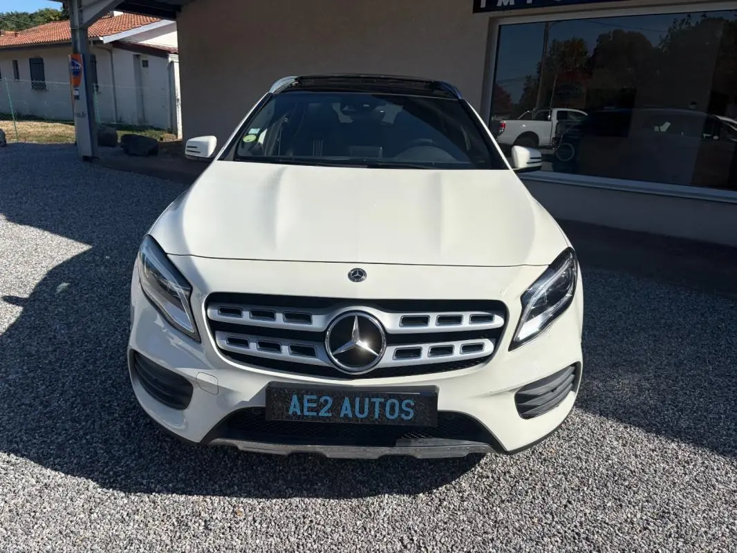 Vue avant d'un Mercedes GLA 220d blanc avec calandre chromée et toit panoramique noir, stationné sur gravier devant un bâtiment.