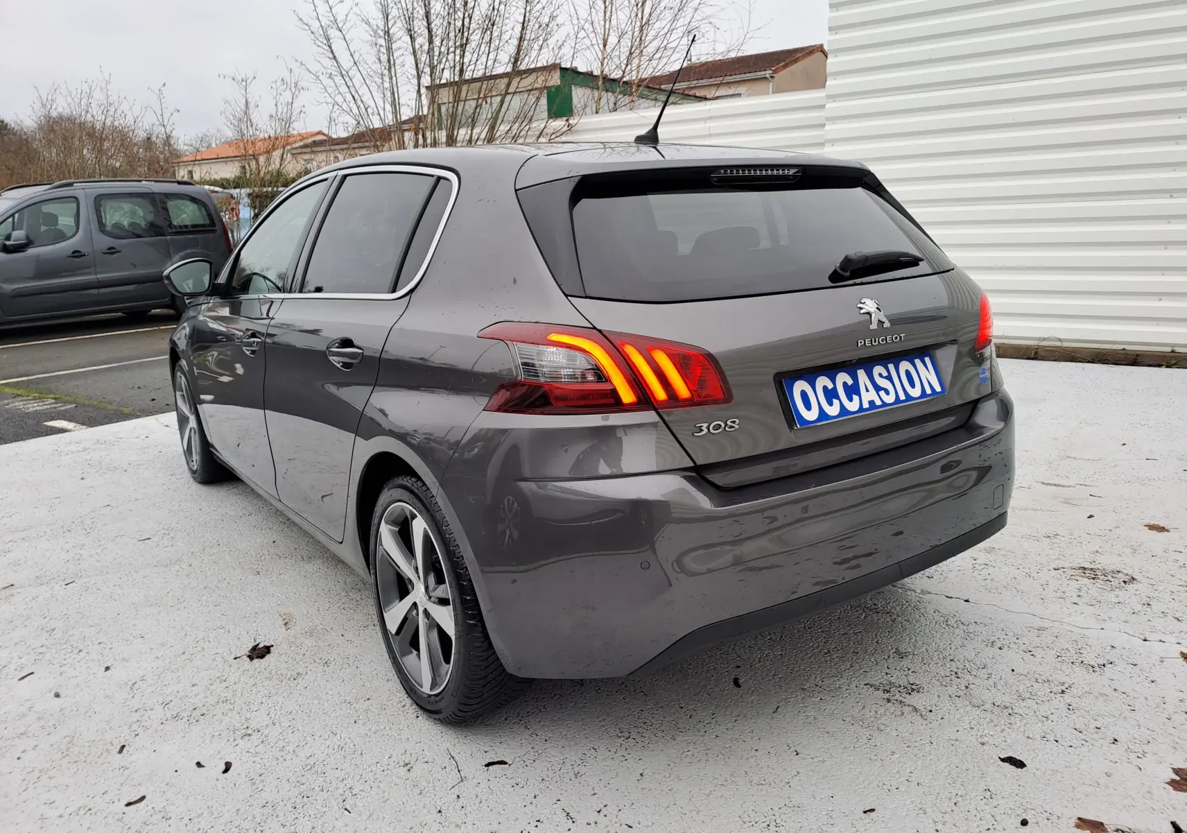 Vue 3/4 arrière droite d'une Peugeot 308 gris métallisé avec feux arrière allumés et plaque "OCCASION" visible.