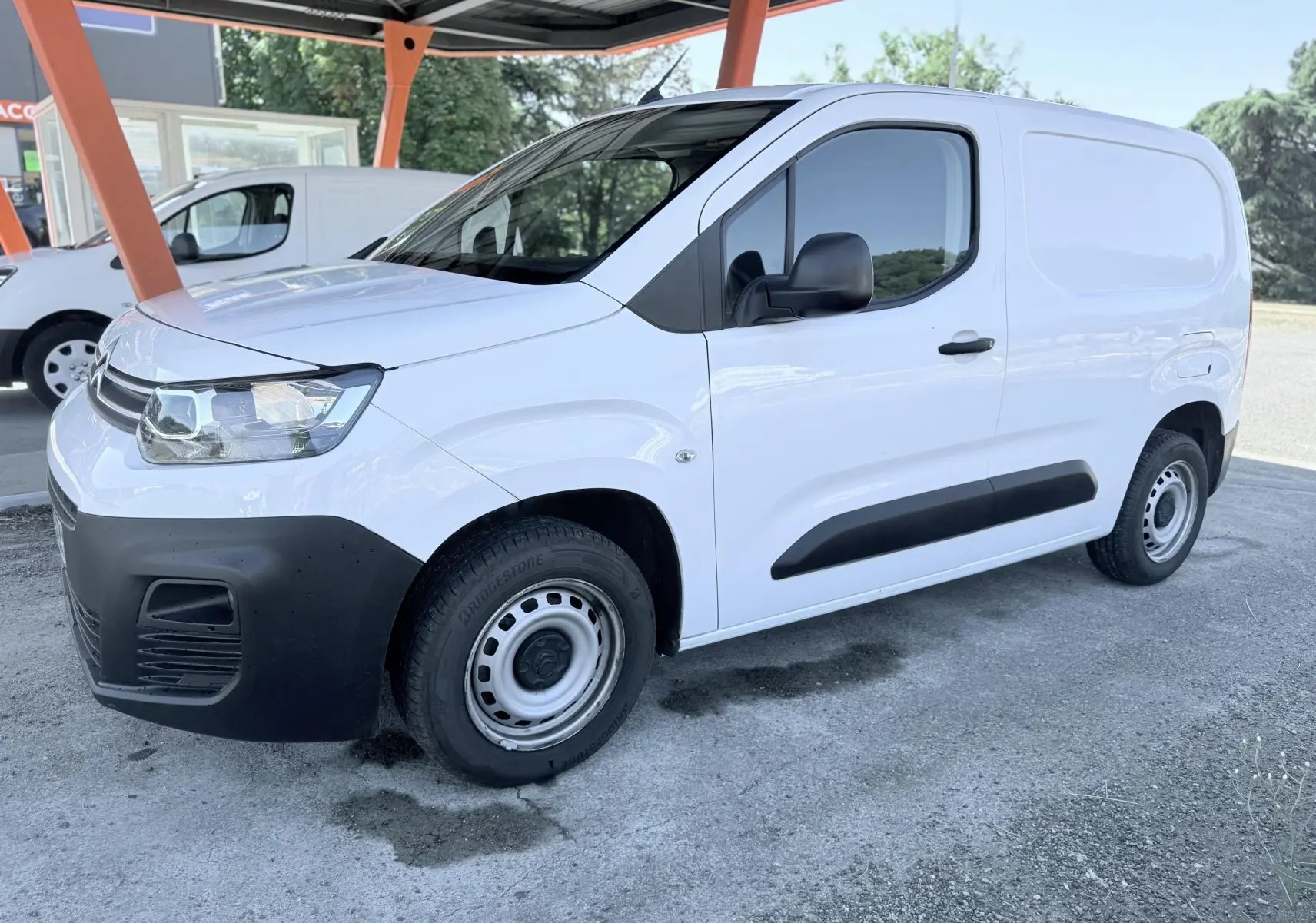 Vue 3/4 avant droit d’un utilitaire Citroën Berlingo blanc avec pare-chocs noirs et jantes acier sous abri.