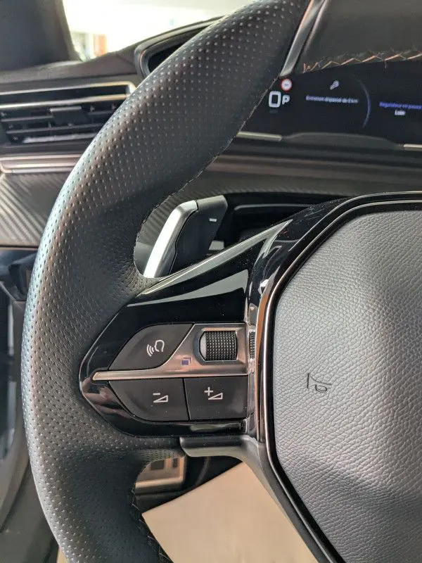 Gros plan sur le volant cuir noir perforé de la Peugeot 508 SW 2024, avec commandes audio intégrées et tableau de bord numérique en arrière-plan.