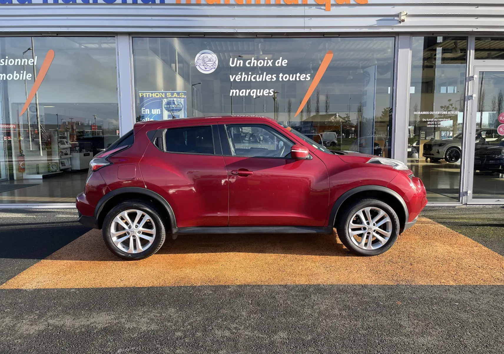 Vue de profil côté gauche d'un Nissan Juke rouge 2017 garé devant une vitrine de concession multimarque.