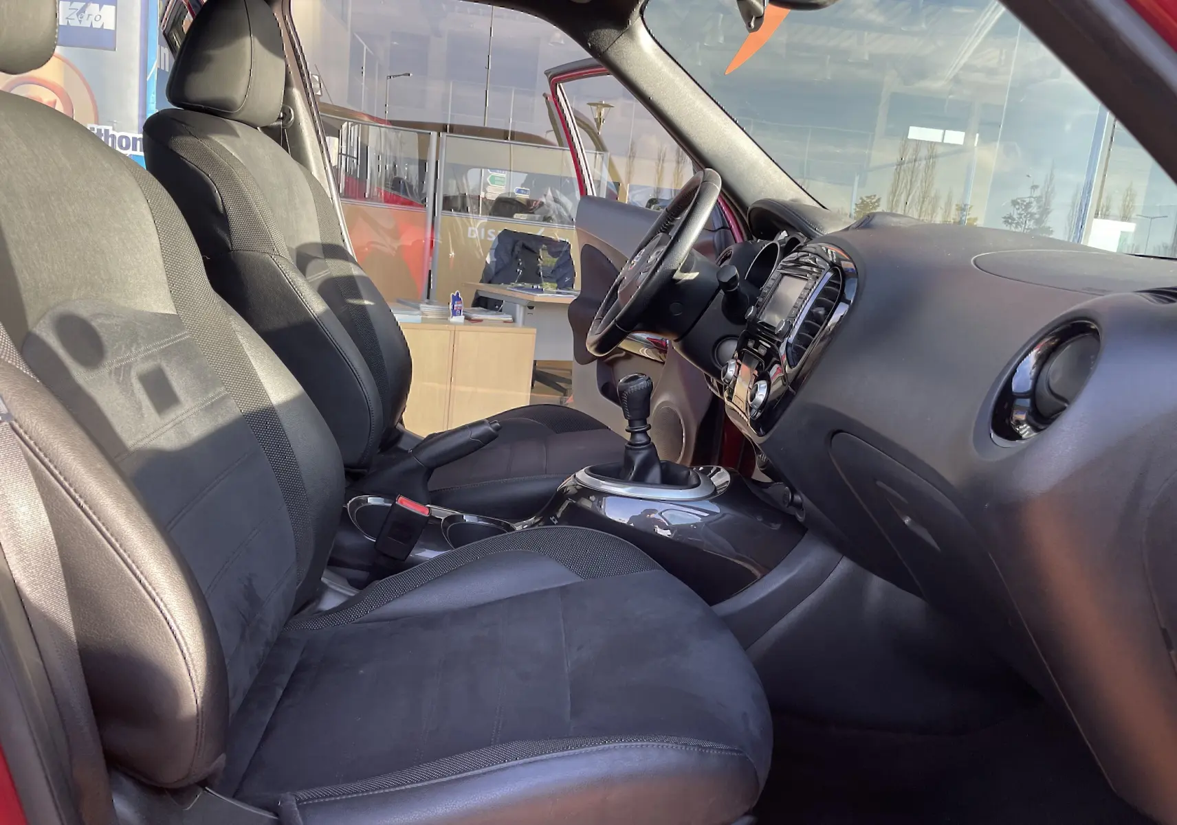 Vue intérieure côté conducteur du Nissan Juke rouge 2017, mettant en valeur les sièges noirs et la console avec levier de vitesse manuel.